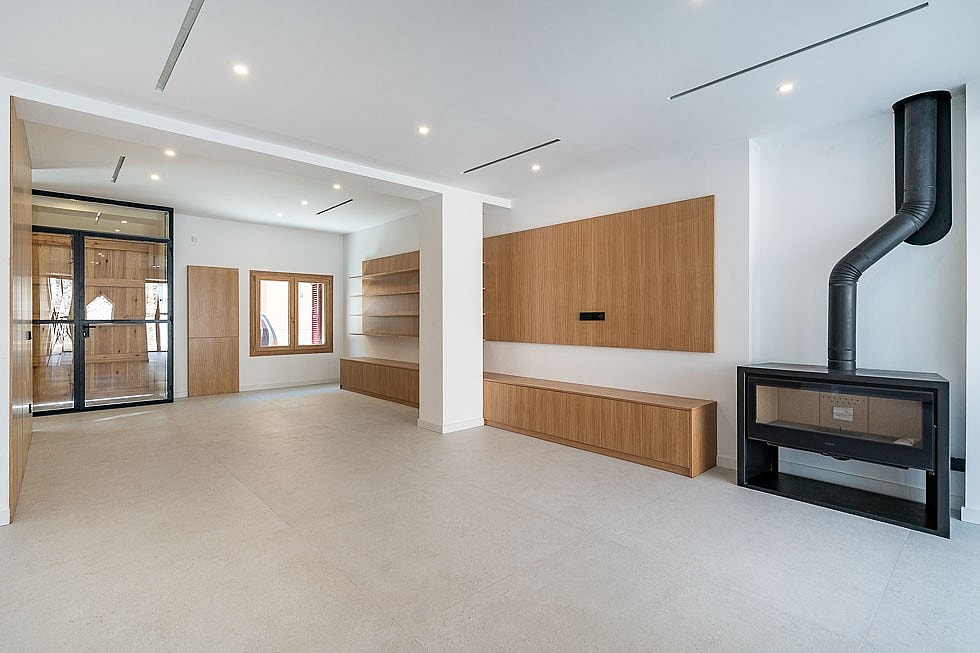 An expansive, bright living space with large light-grey floor tiles, custom light oak cabinetry, and a black metal fireplace.