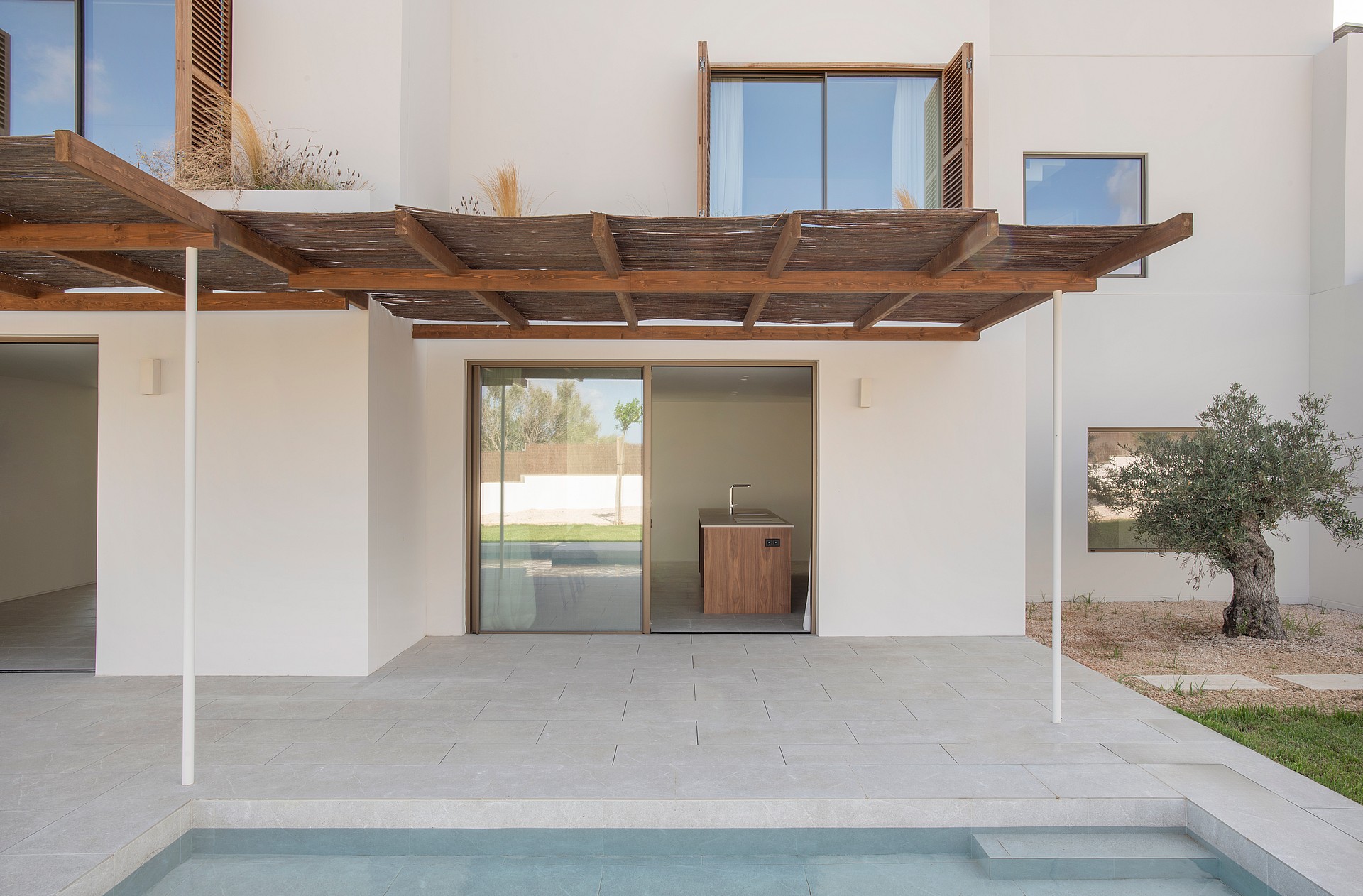 A stone terrace with a wooden pergola sits beside a swimming pool, leading to a modern white villa with glass doors.