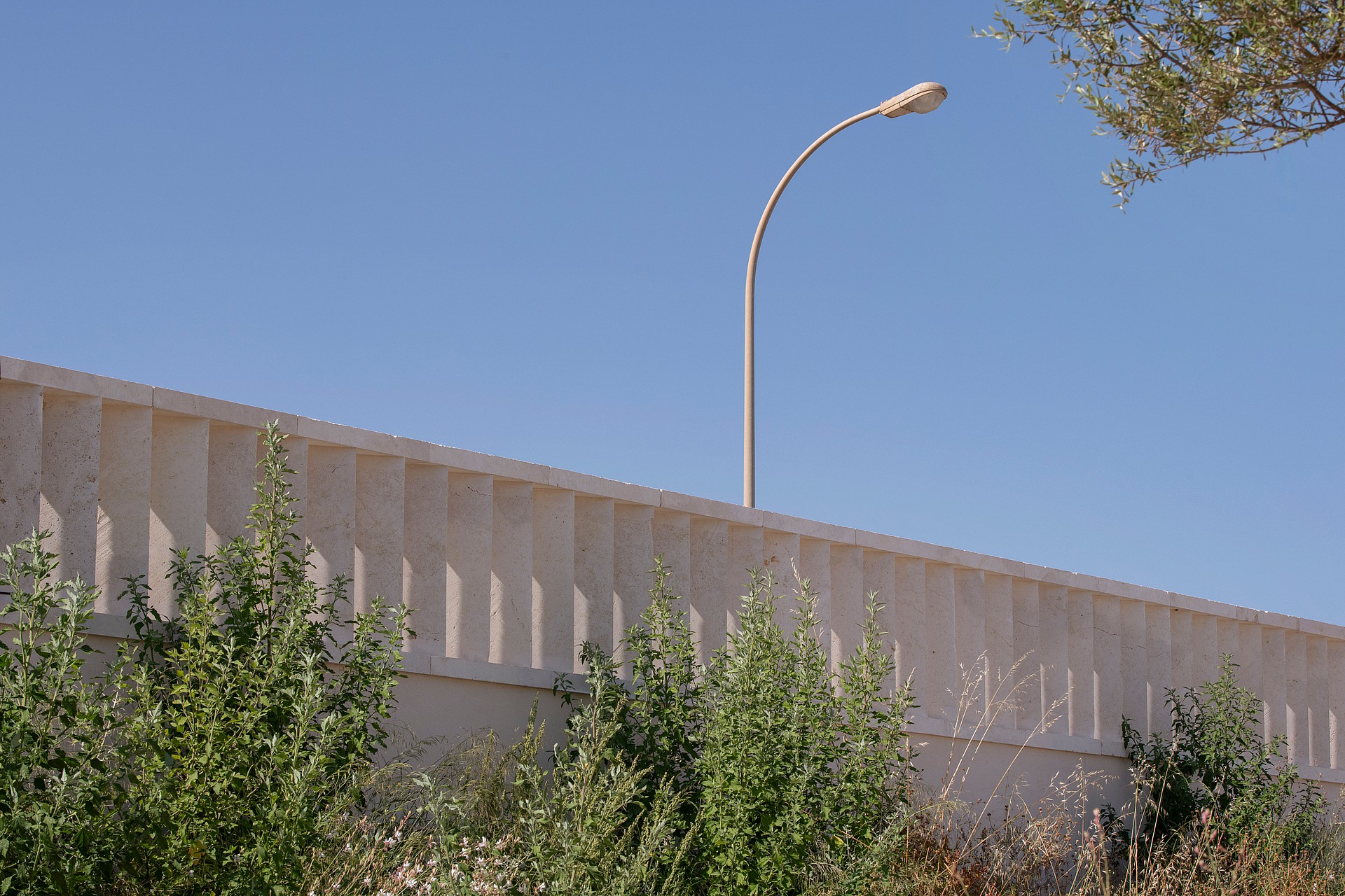 A beige stone wall featuring vertical recessed slats stands under a clear blue sky with a curved metal street lamp.