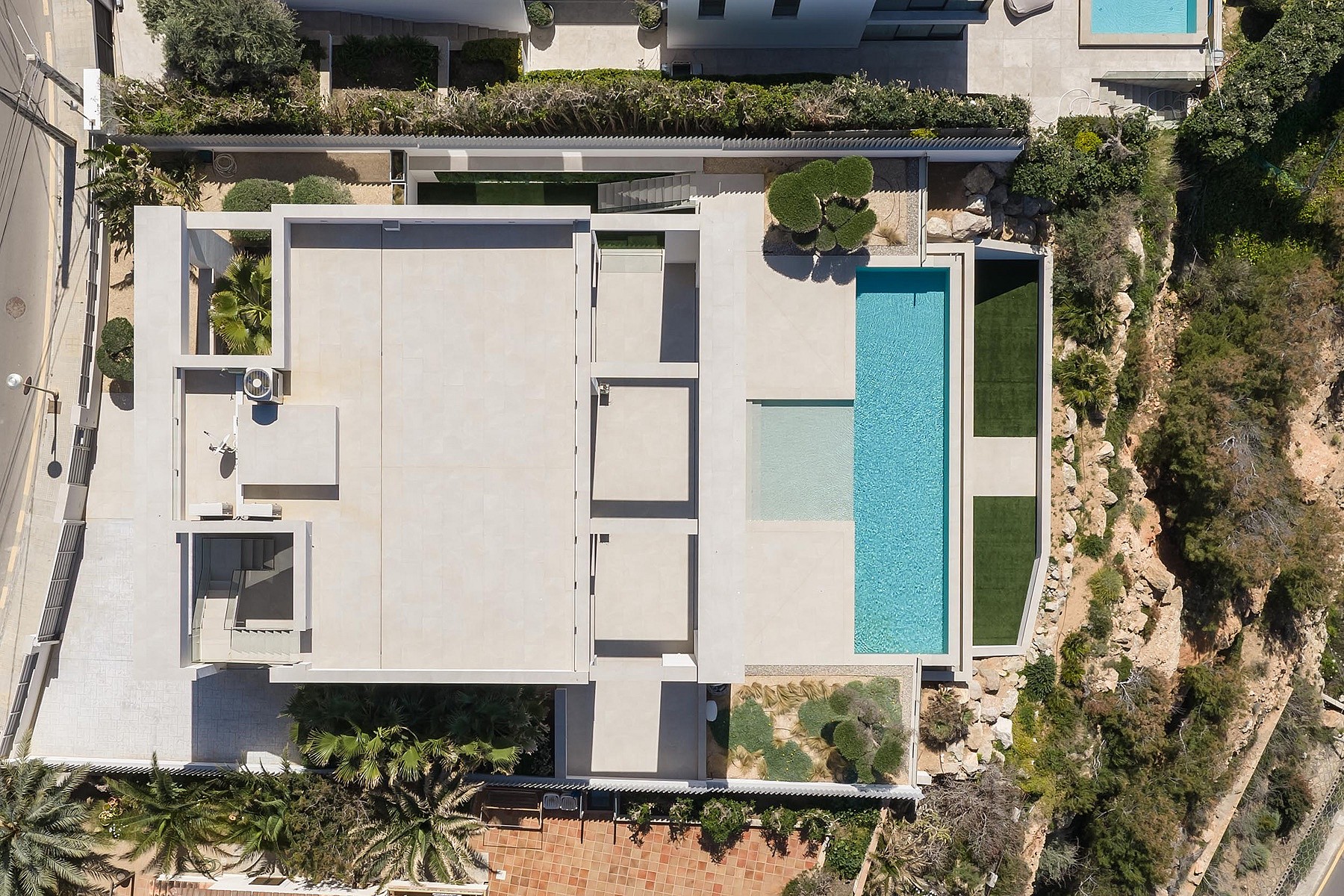 Top-down drone shot of a modern white villa with a rectangular swimming pool, terraced gardens, and surrounding Mediterranean landscape.