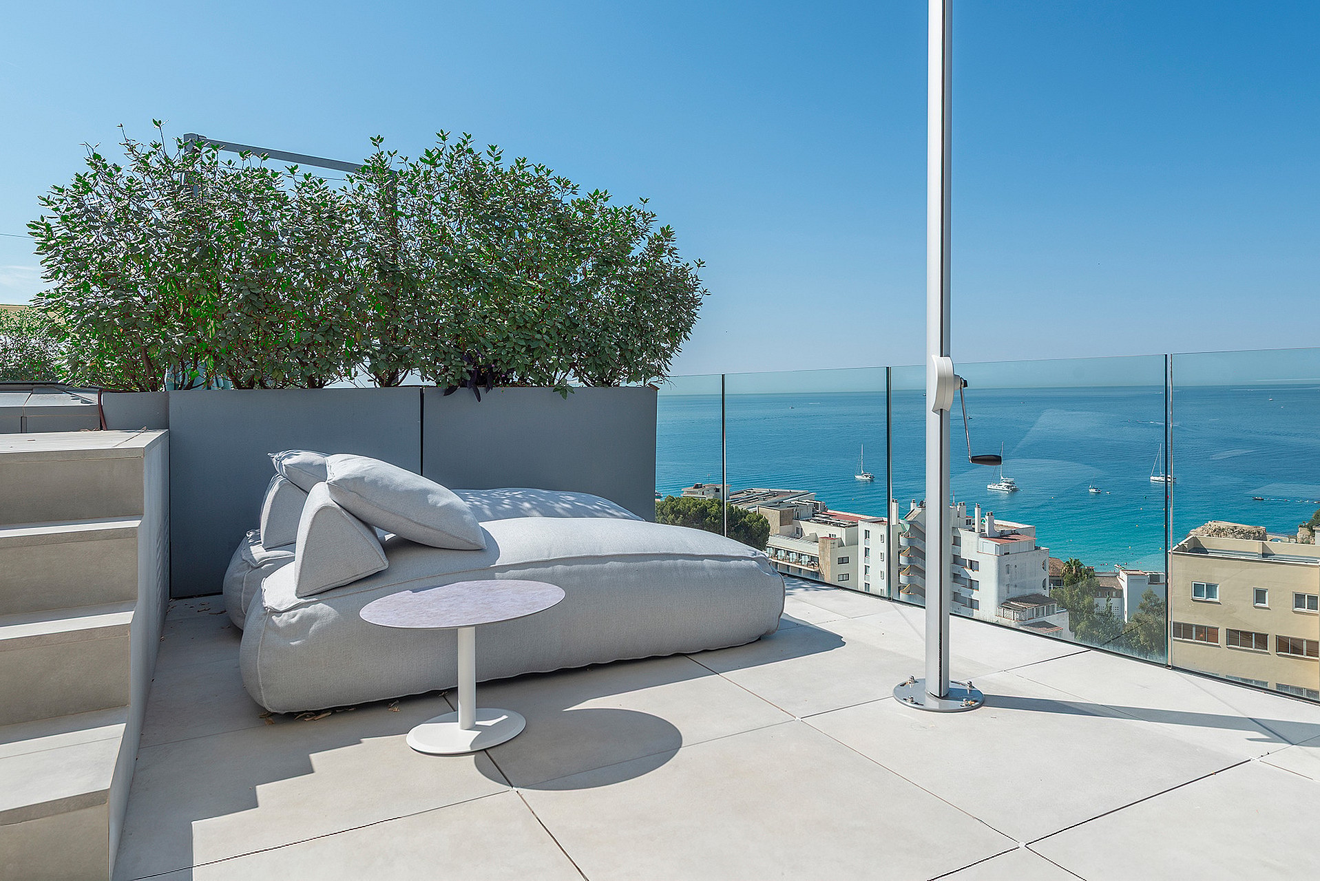 A rooftop terrace featuring a large gray outdoor daybed, a white side table, and a glass balustrade overlooking the sea.