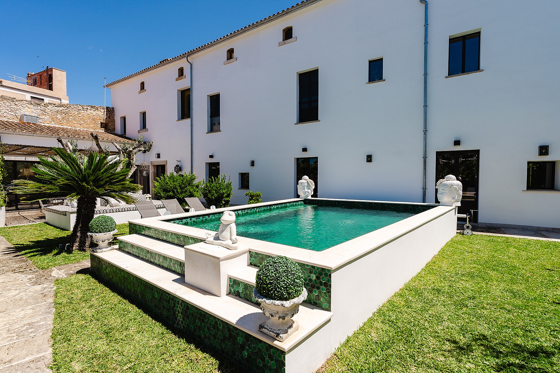 A raised outdoor swimming pool with green tiled edges and stone steps, set in a lawn next to a white traditional townhouse facade.