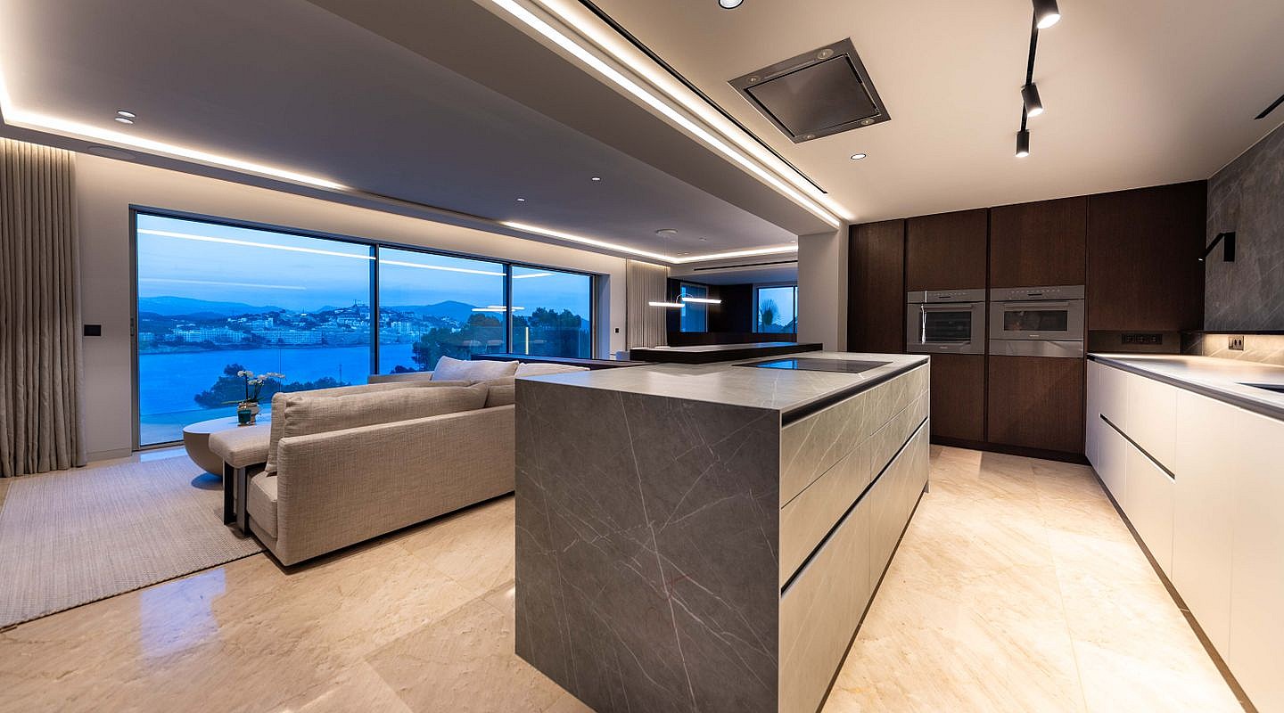 A contemporary kitchen featuring a stone-topped central island, dark wood cabinetry, and integrated appliances under recessed lighting.
