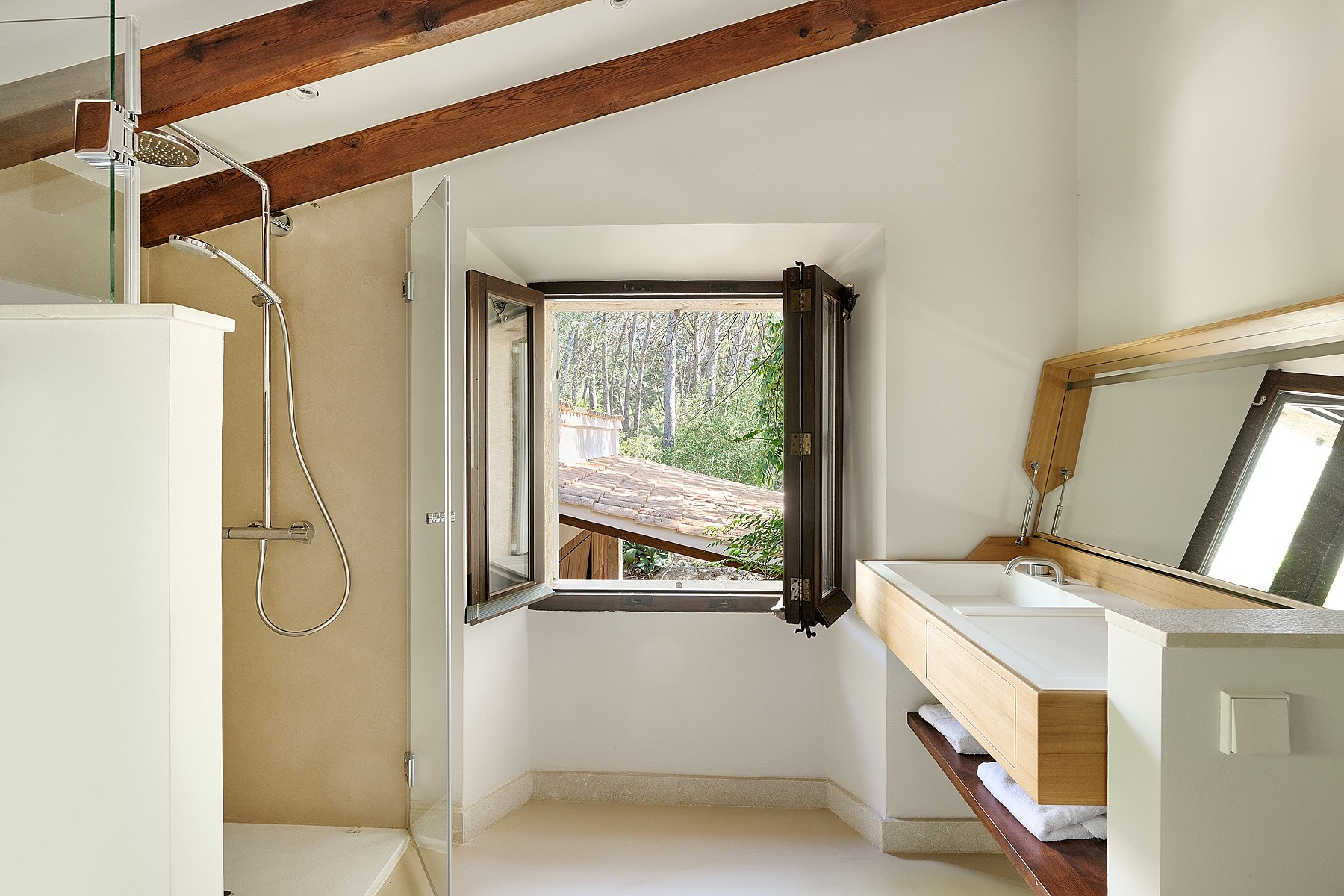 A bathroom in a finca with white walls, wooden ceiling beams, a floating vanity, and an open window showing trees.