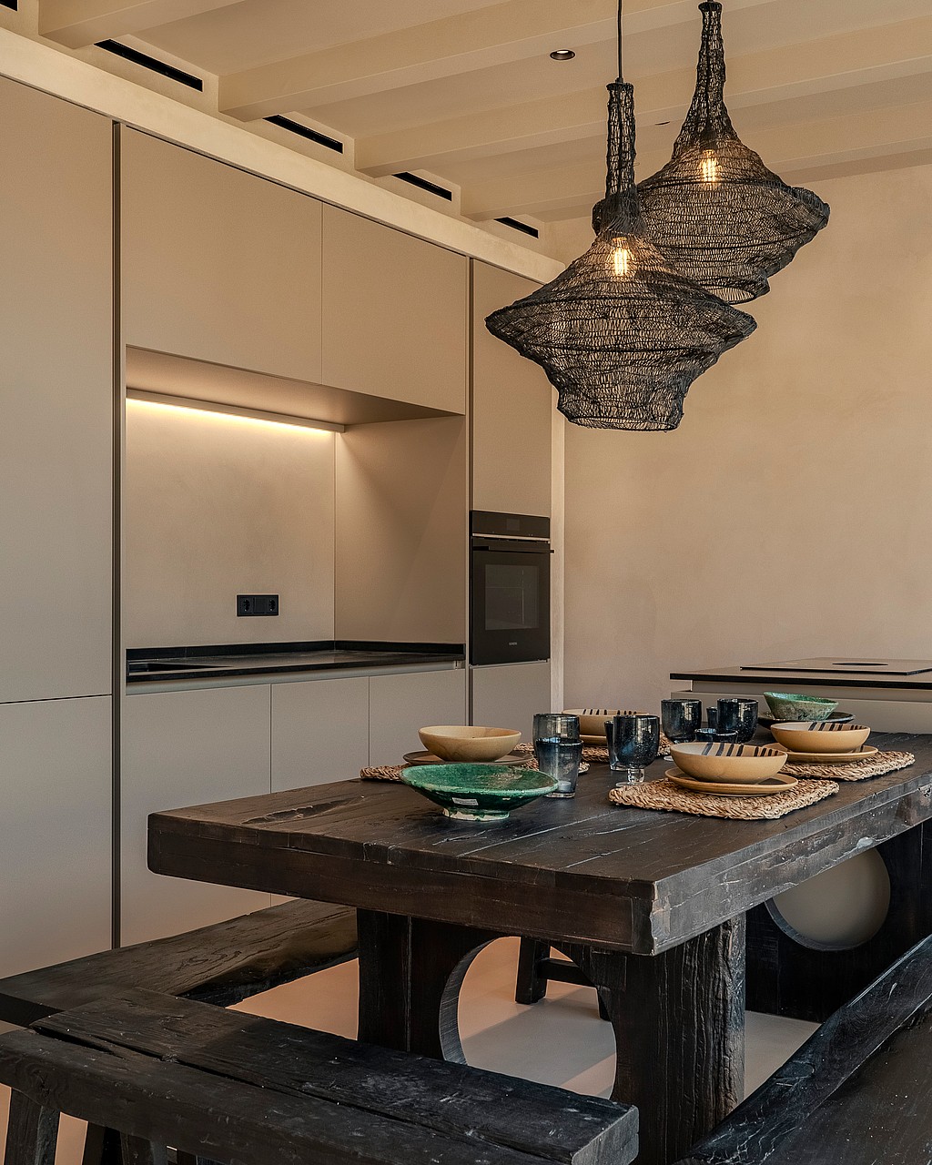 A modern kitchen featuring floor-to-ceiling beige cabinets and a dark, weathered timber dining table with matching bench seating.