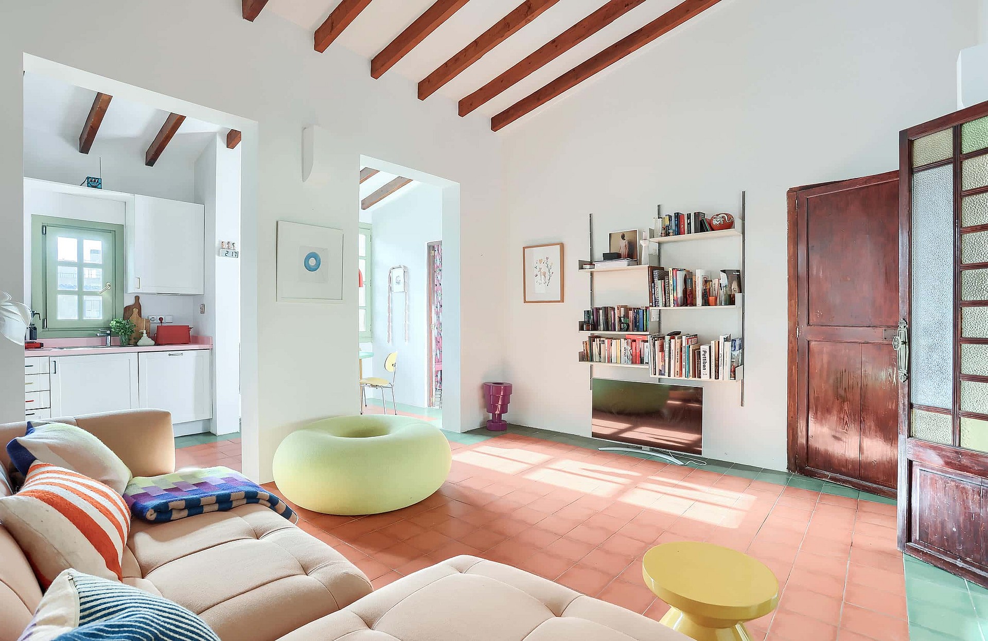 A bright living room with terracotta tiled floors, exposed dark wooden ceiling beams, and white walls. A large beige sofa sits in the foreground, with a circular lime green ottoman in the center and open shelving against the wall.
