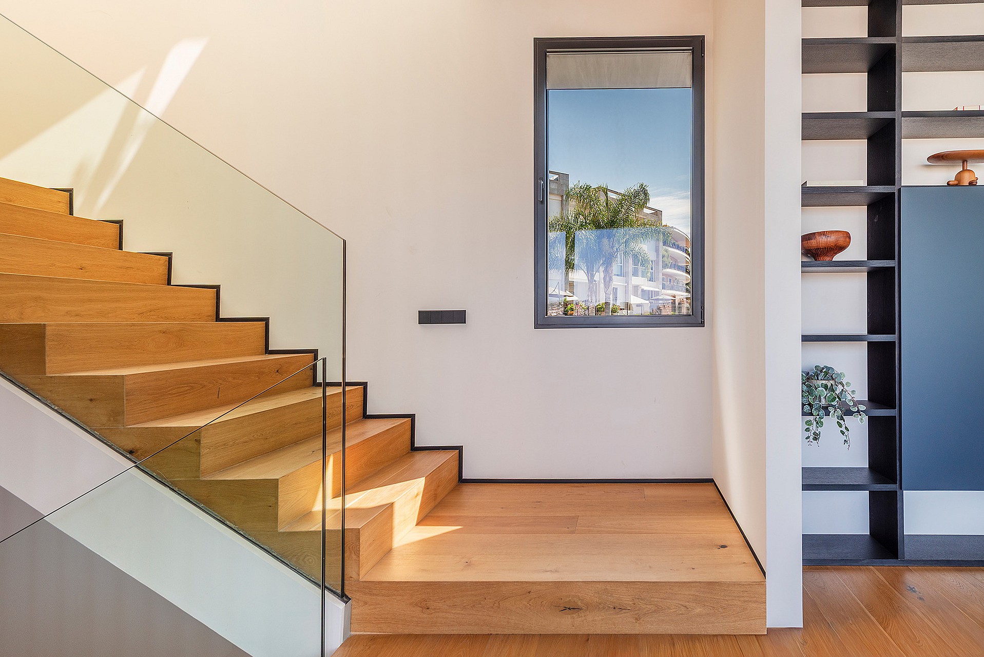 Interior wooden staircase with glass railing, light oak treads, and a built-in dark shelving unit beside a square window.