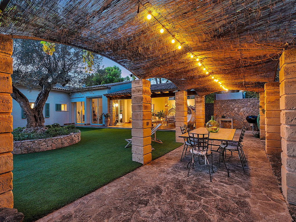 A covered terrace with natural stone pillars and a reed roof, featuring an outdoor dining area with wrought iron chairs, overlooking a landscaped garden with an olive tree at dusk.