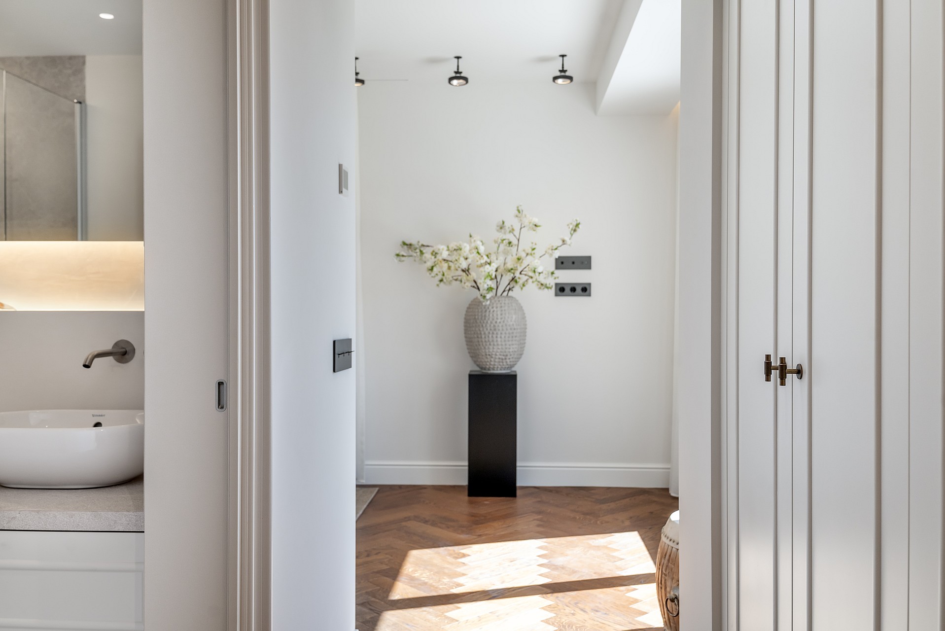 A view through a doorway showing herringbone oak floors, white walls, and a black pedestal with a floral arrangement.