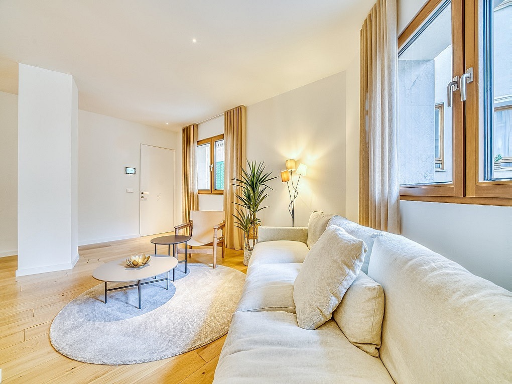 A modern living room with light-colored sofa, round area rug, wooden coffee table, and large windows looking onto the city.