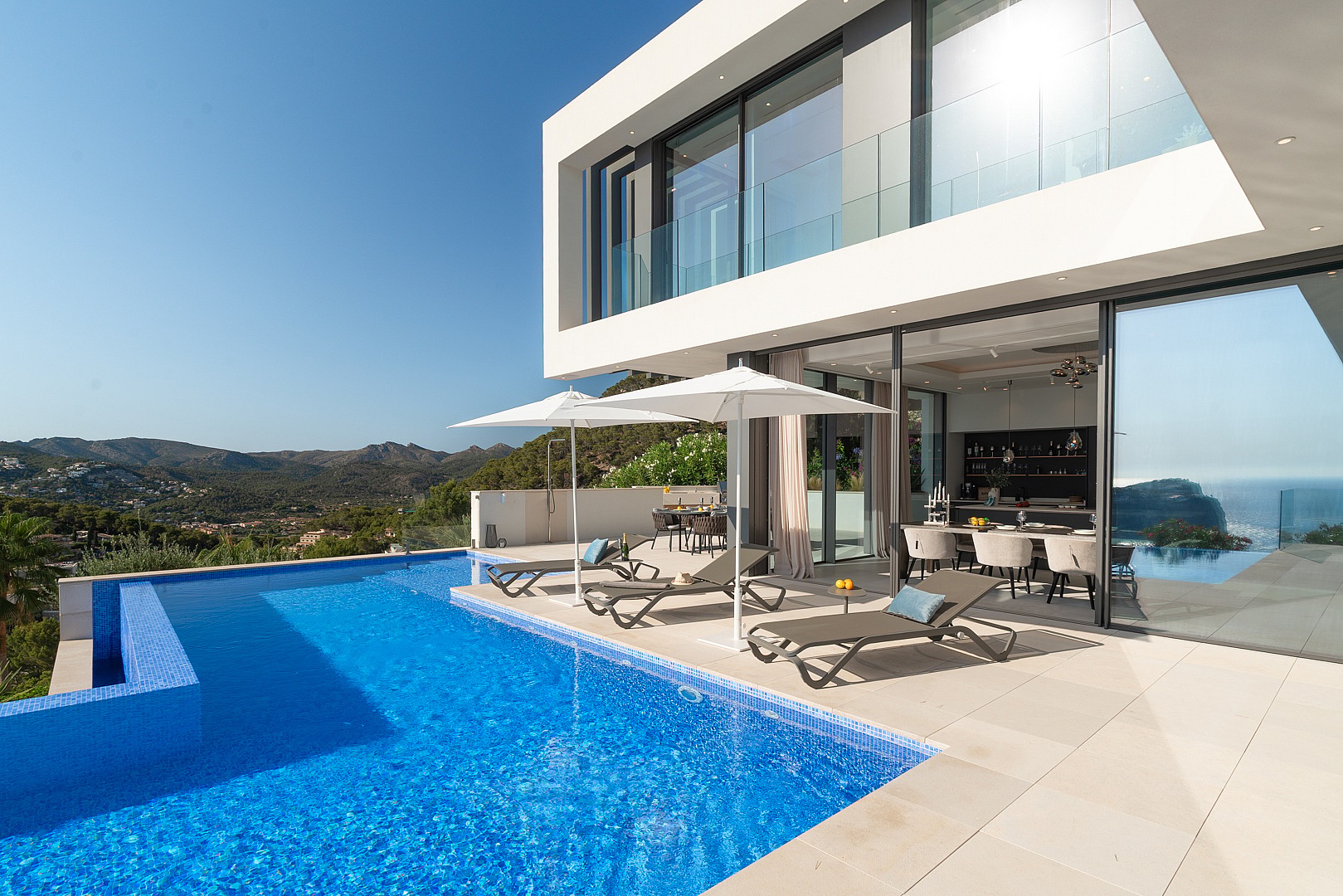 Modern villa with a blue tiled infinity pool and sun loungers on a stone terrace, set against a Mediterranean coastal landscape.