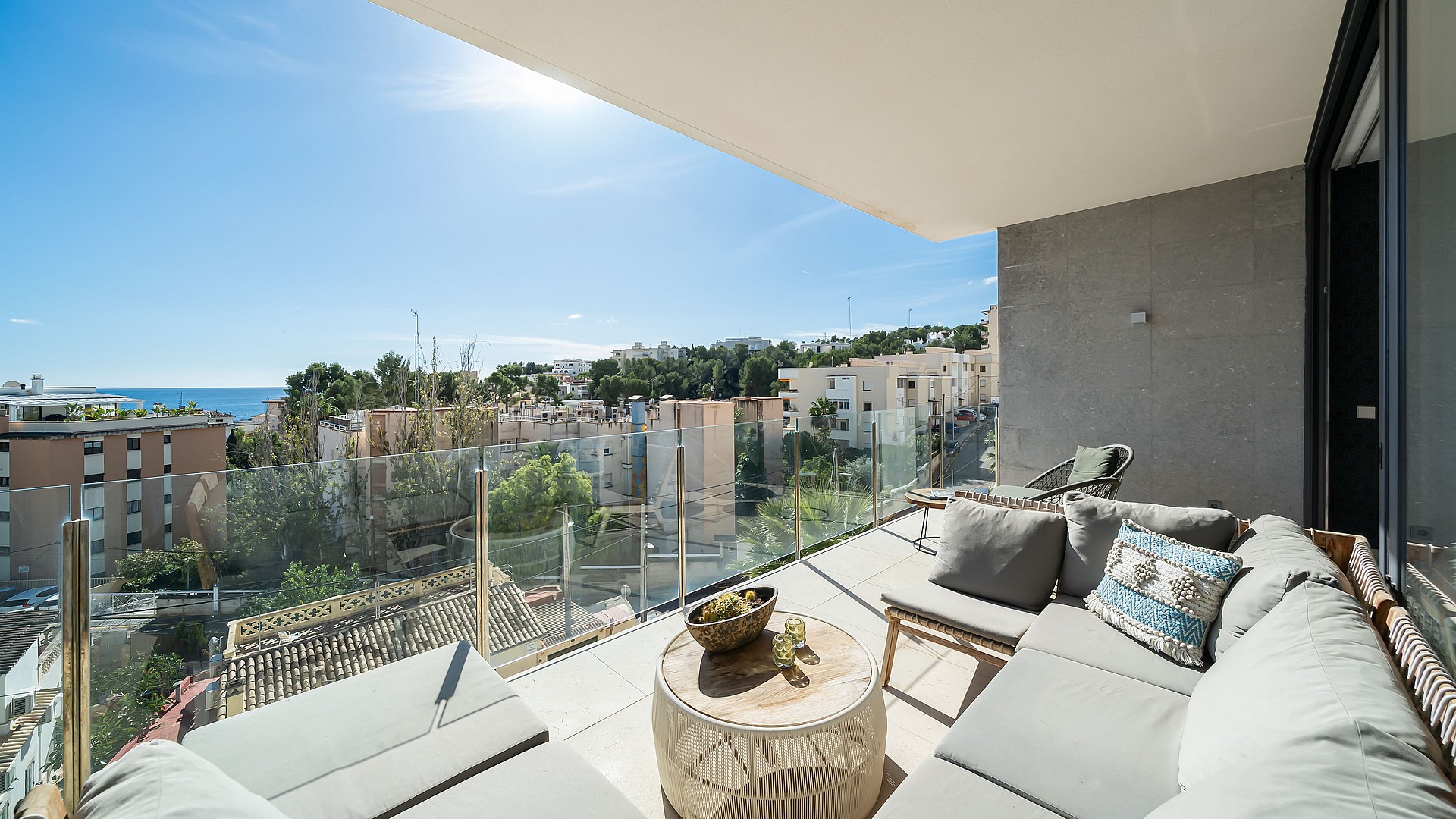 A covered terrace with grey outdoor sofas, a circular wooden coffee table, and glass railings overlooking a coastal town.