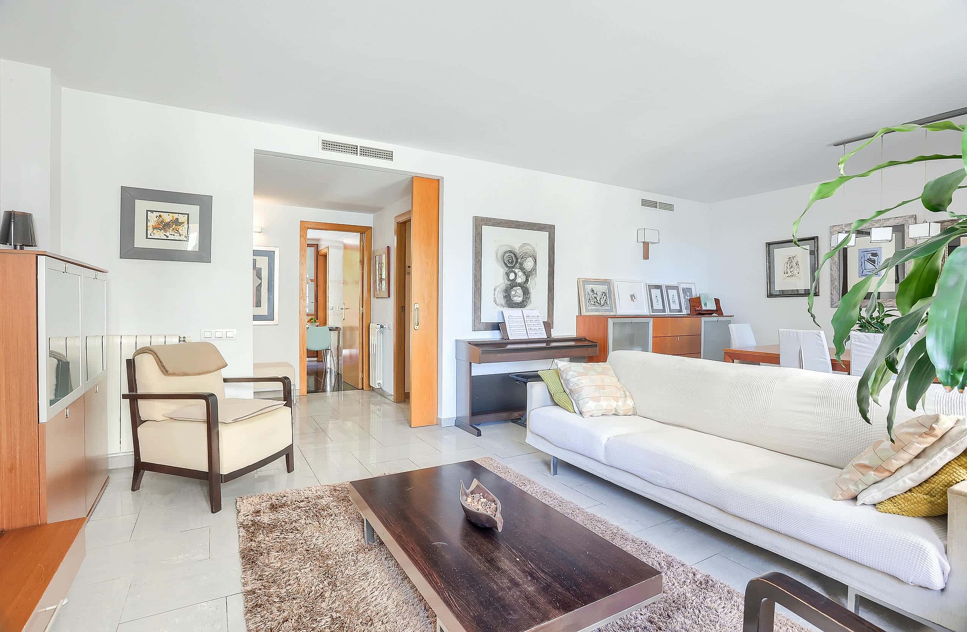 A bright living room with white walls, beige sofa, dark wood coffee table, and an armchair, leading to an interior hallway.