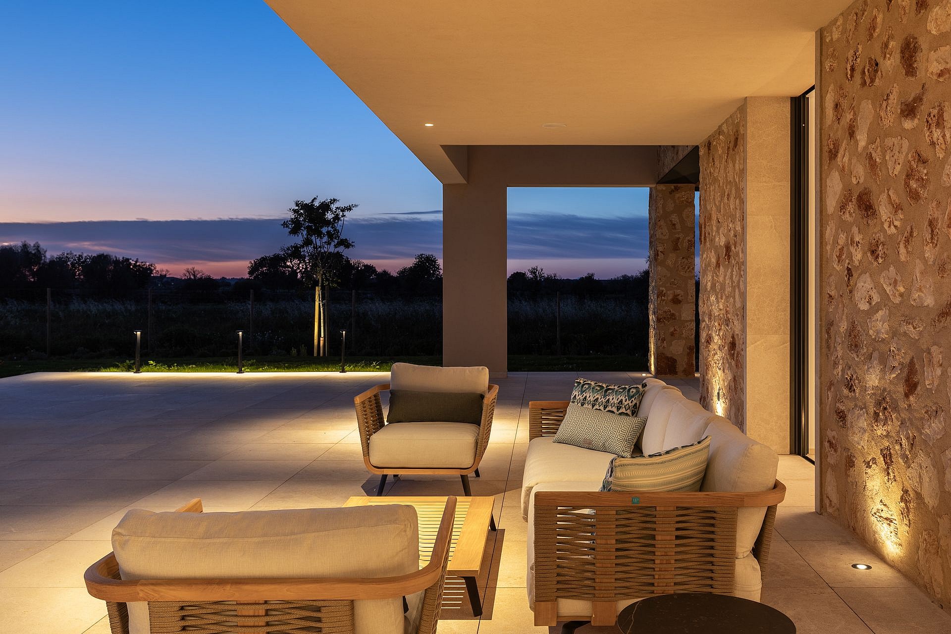 A covered stone terrace featuring woven outdoor seating with soft cushions during twilight, overlooking a dark garden.