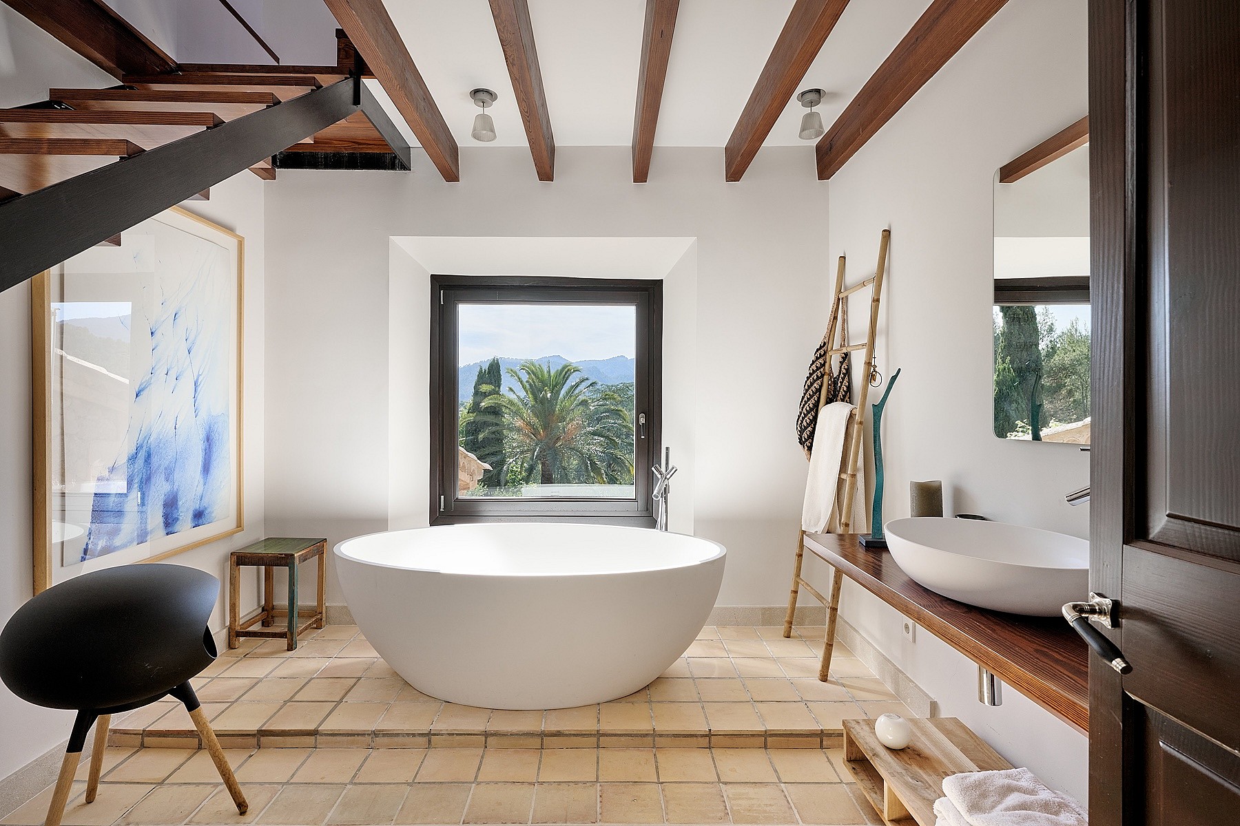 A modern bathroom with a freestanding white soaking tub on a tiled platform, centered before a window with mountain views.