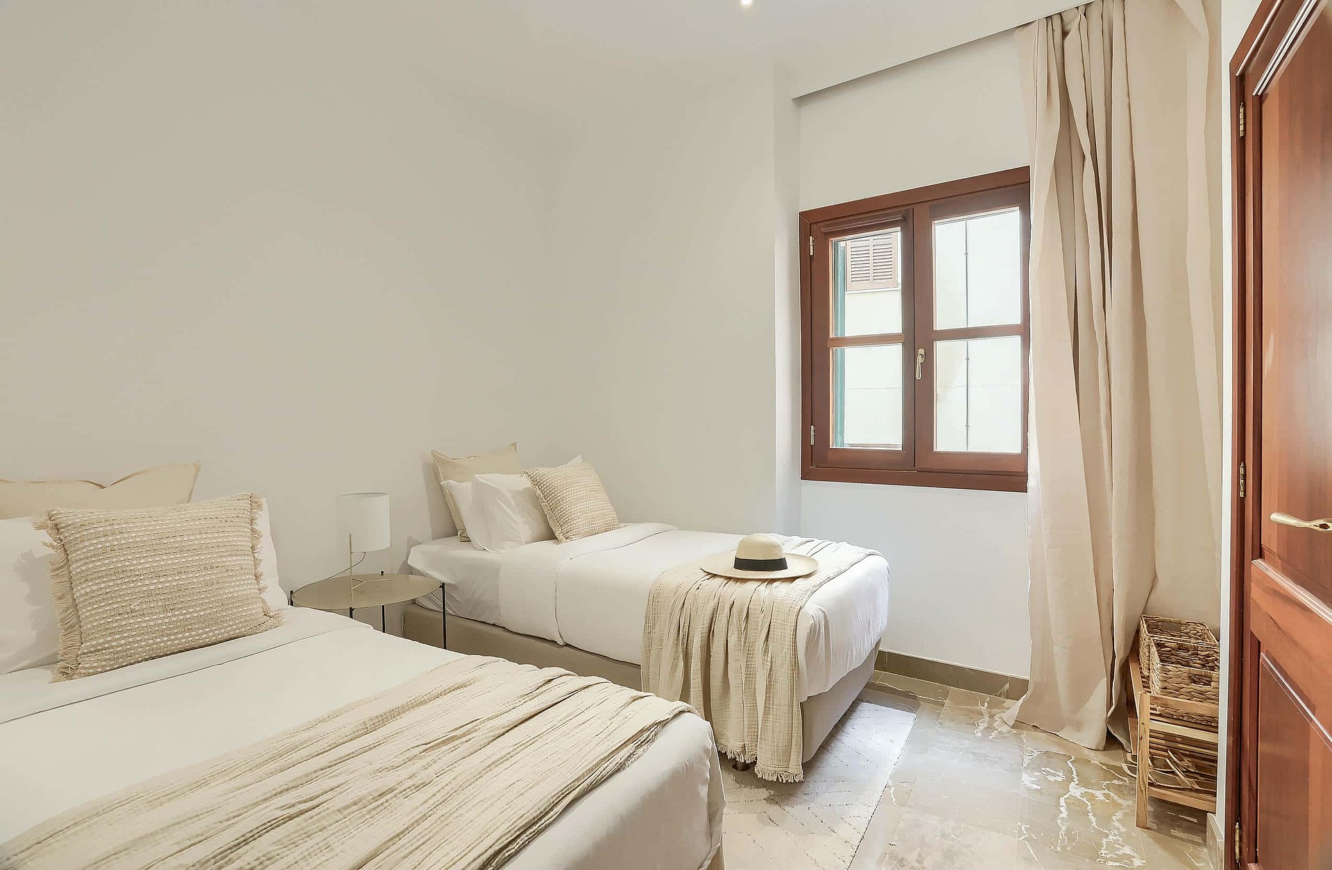A bright bedroom with two twin beds, white walls, light tile flooring, and a wooden window frame with beige curtains.
