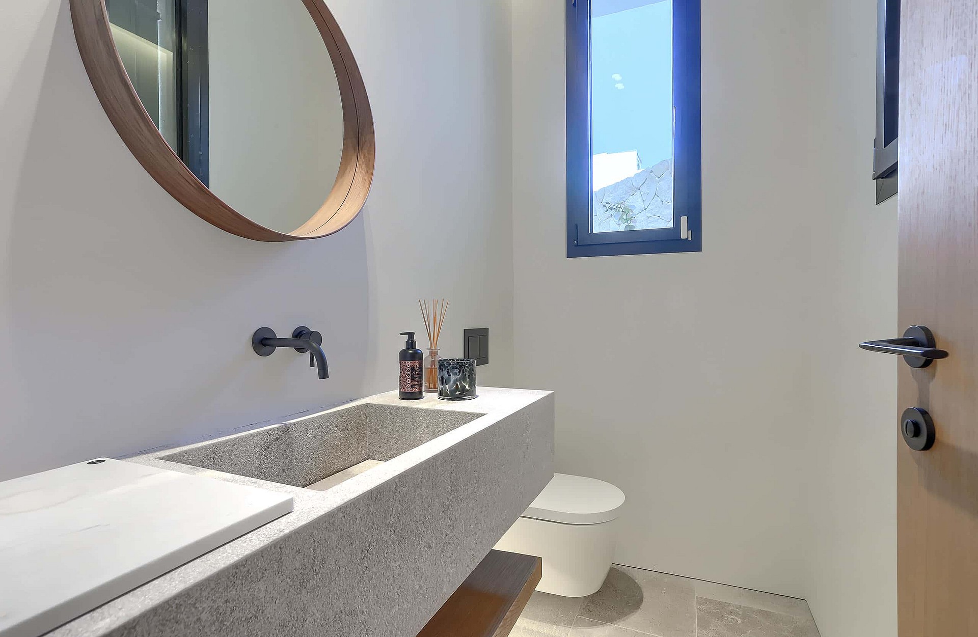 Modern bathroom featuring a textured stone floating vanity, circular wooden-framed mirror, and matte black wall-mounted faucet.
