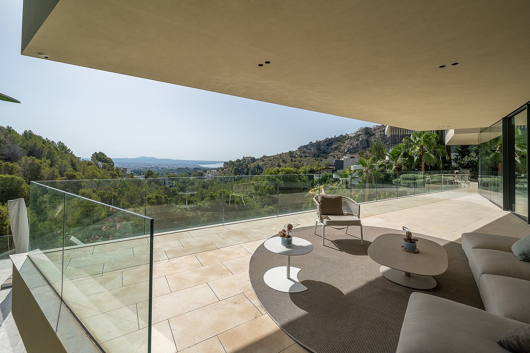 A wide, covered outdoor terrace overlooking hills and a distant coastline, featuring neutral outdoor furniture, glass railings, and stone flooring.