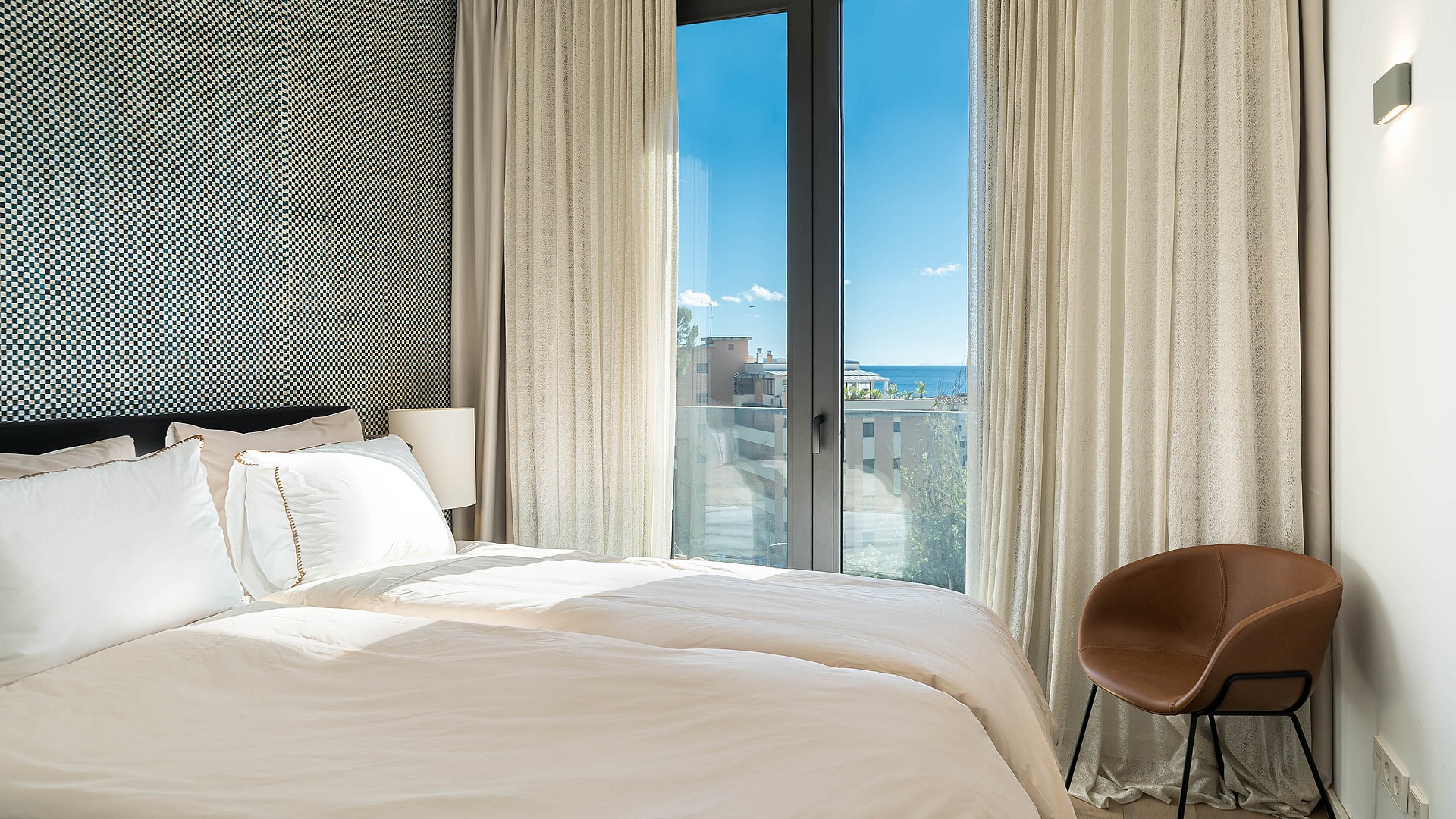 A bedroom corner featuring a black and white geometric patterned wallpaper, white bedding, and floor-to-ceiling glass doors.