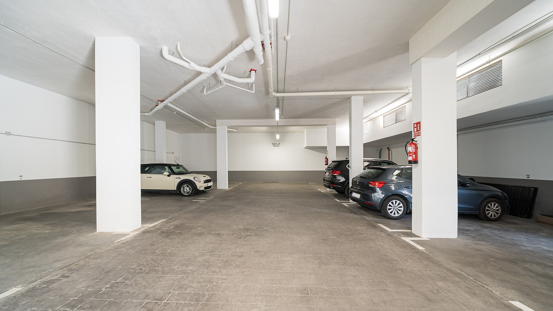 A clean, well-lit underground parking garage with concrete floors, white painted columns, and two parked cars.