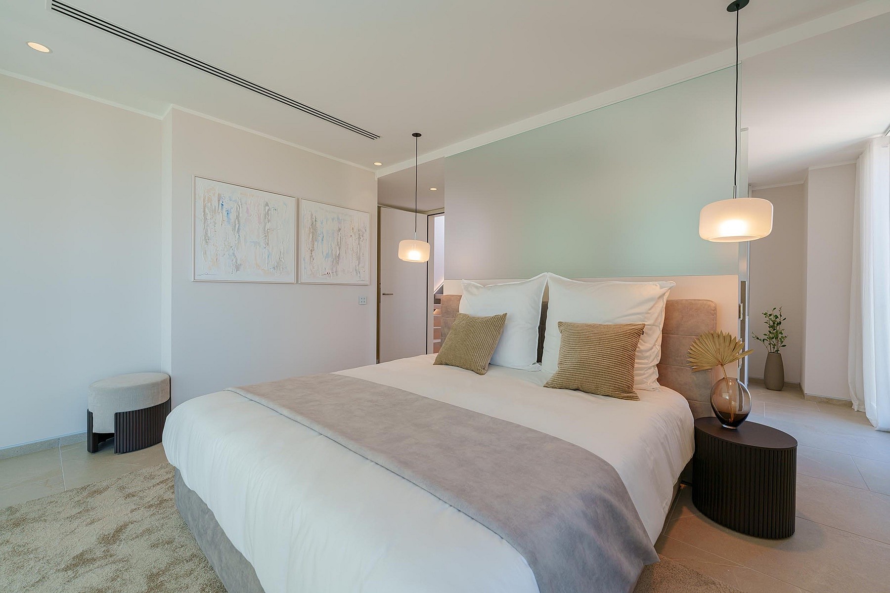 Dormitorio luminoso con cama de matrimonio, ropa de cama blanca, cojines neutros e iluminación colgante.
