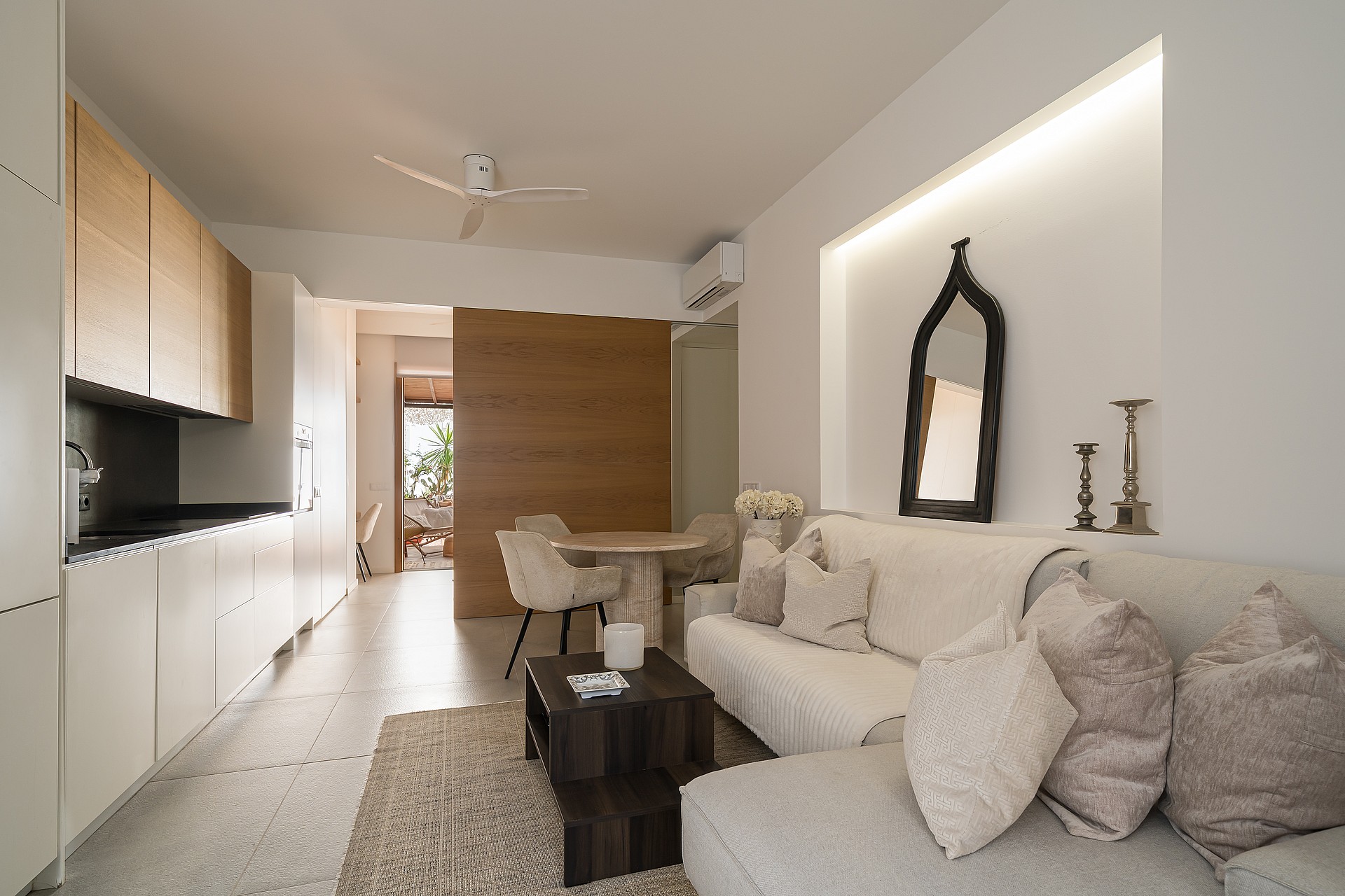 A bright apartment living space featuring a white sofa, minimalist kitchenette with wood cabinets, and dining area.