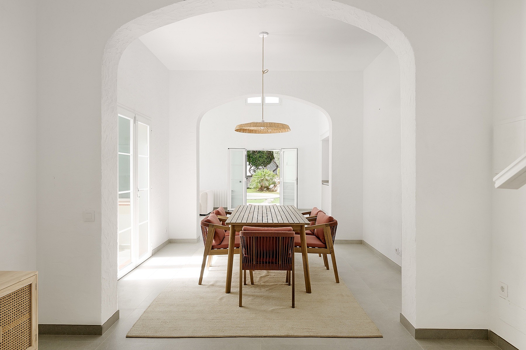 Comedor con bóveda blanca, mesa de teca, sillas de cuerda y lámpara de ratán, vistas al jardín a través de puertas abiertas