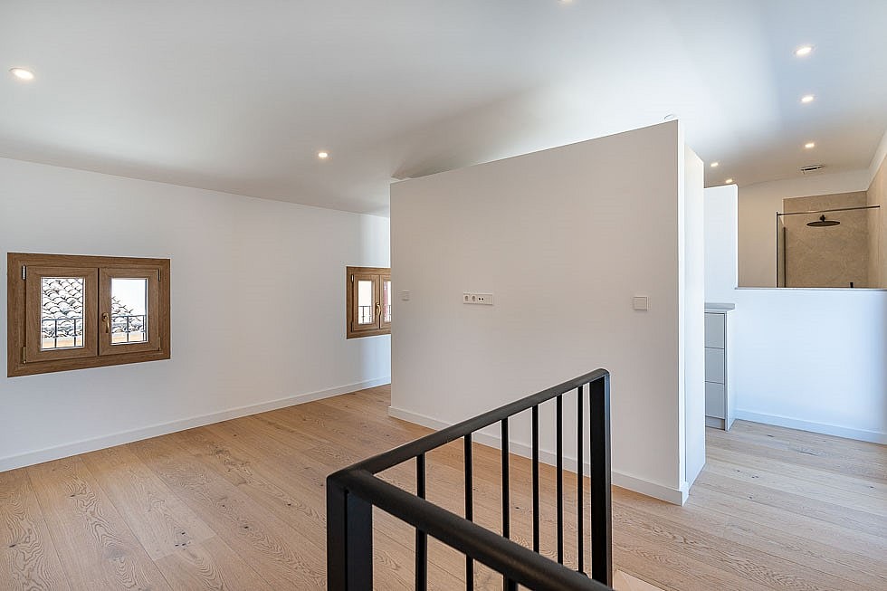 An open landing area in a renovated townhouse with white walls, light wood flooring, a black metal staircase railing, and wooden window frames.