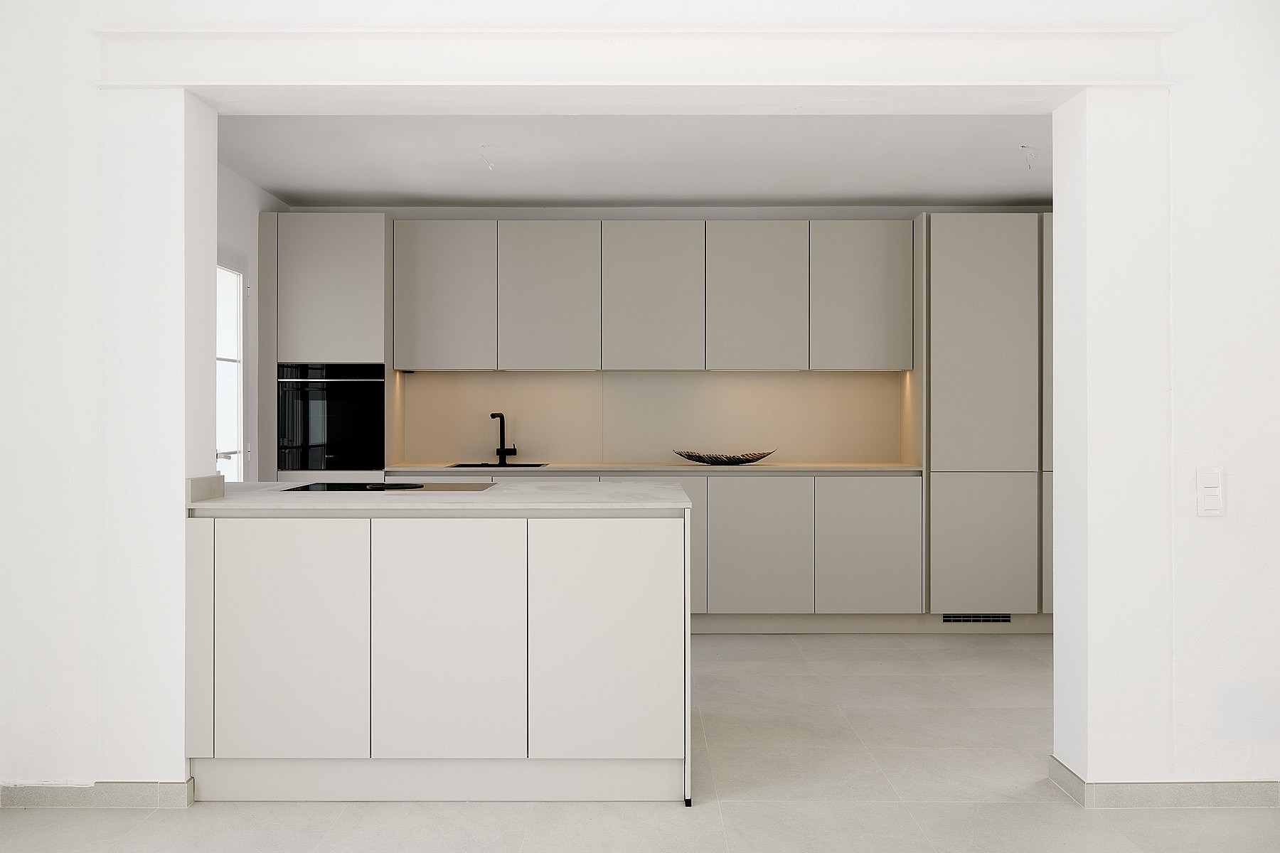 Cocina taupe mate con isla central, encimeras de piedra compuesta y grifería negra vista a través de una abertura blanca