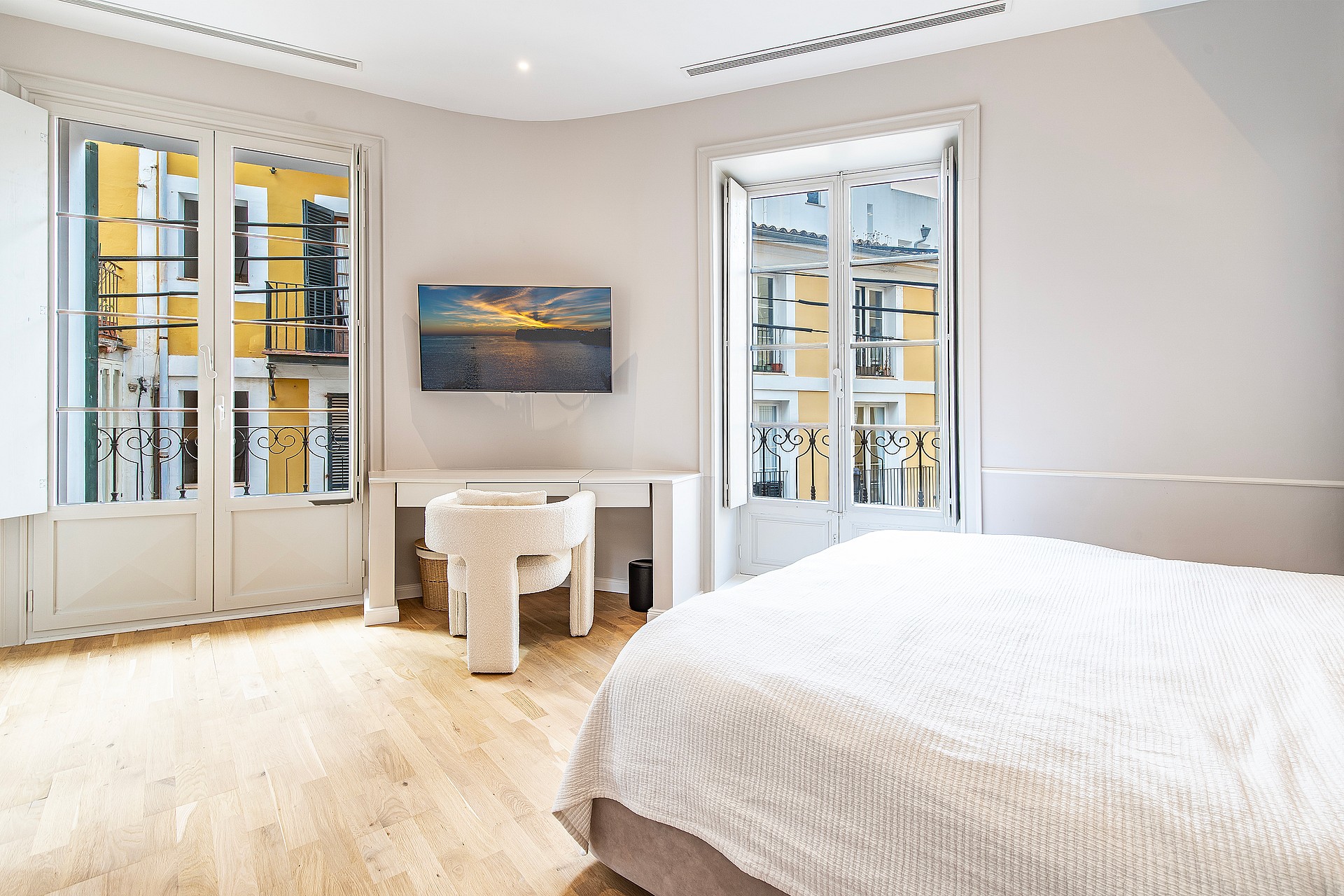 Bright bedroom with white walls, light wood floors, a desk, and floor-to-ceiling windows looking out onto a yellow building.