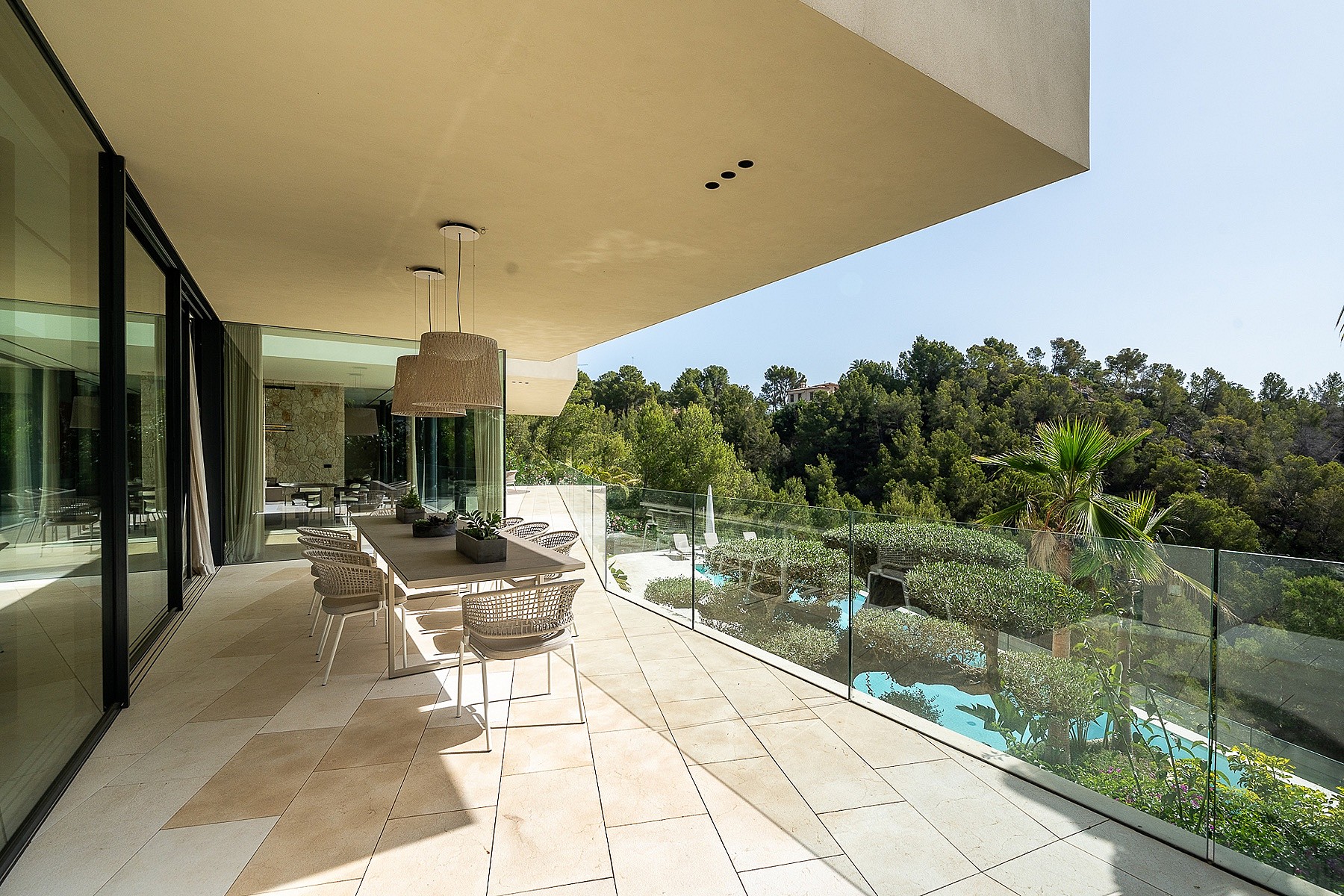 An expansive covered outdoor terrace featuring a dining table, natural stone flooring, and a glass balustrade overlooking trees.