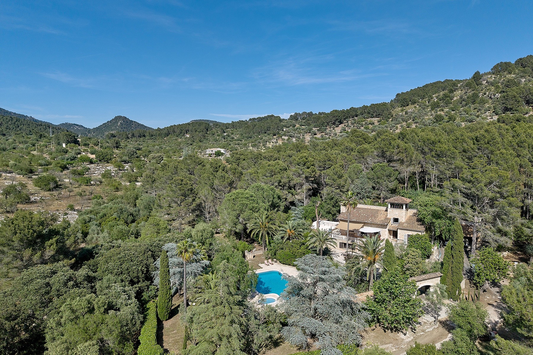 Aerial shot of a stone finca surrounded by dense green forest and mountains under a clear blue sky, with a visible pool.