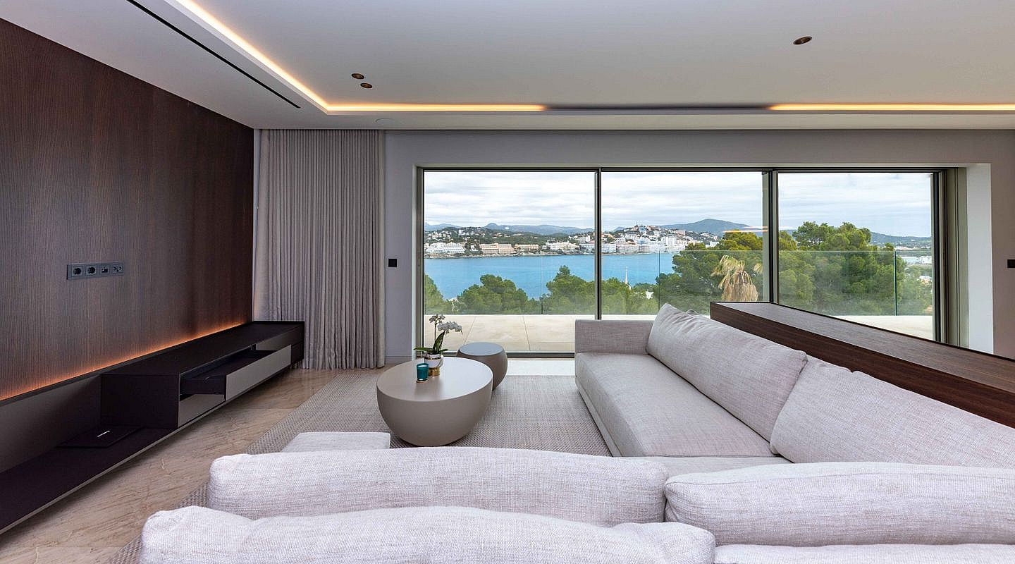 Modern living room with a long beige fabric sectional sofa facing floor-to-ceiling glass windows overlooking a blue bay.