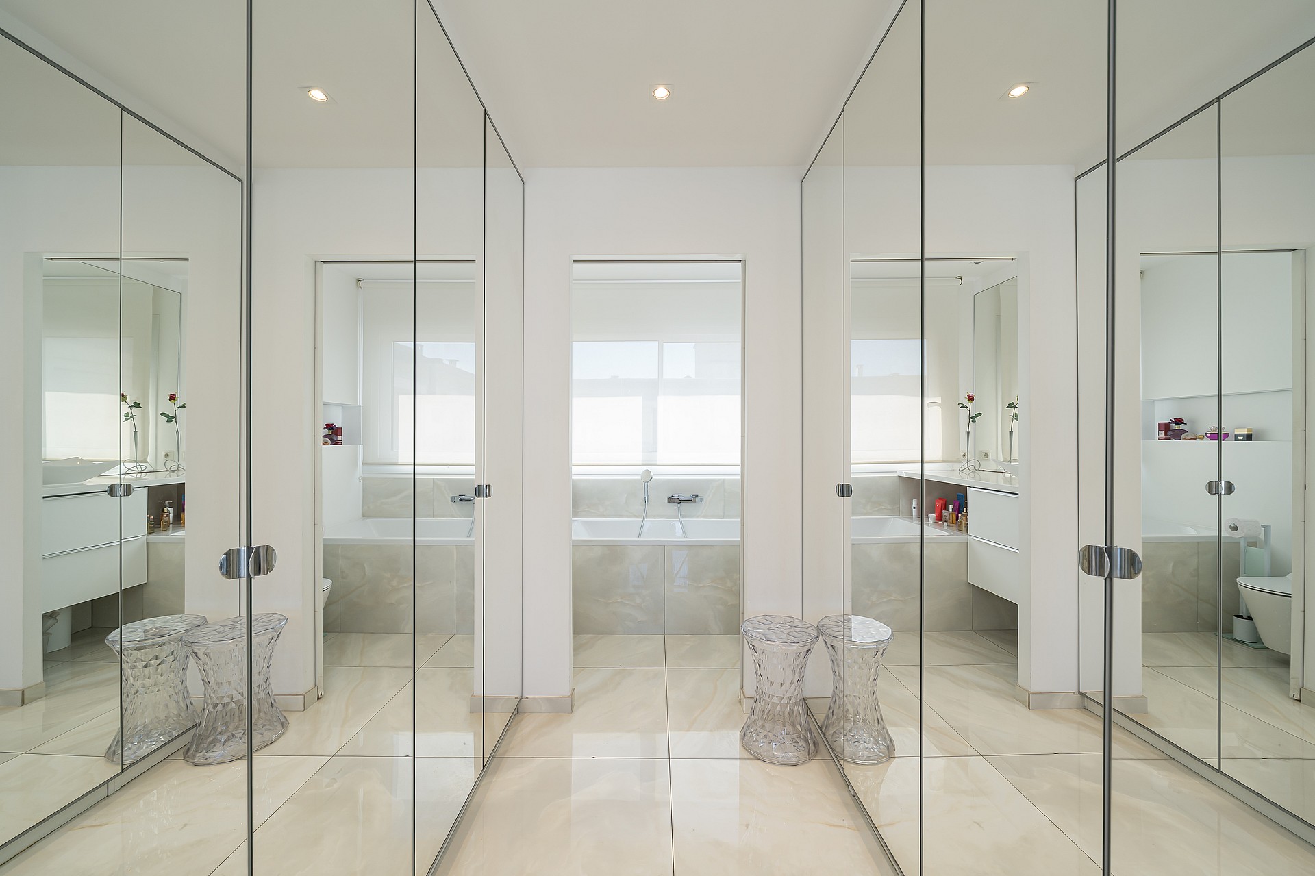 A bright dressing room featuring wall-to-wall mirrored wardrobes with chrome handles, leading into a white tiled bathroom.