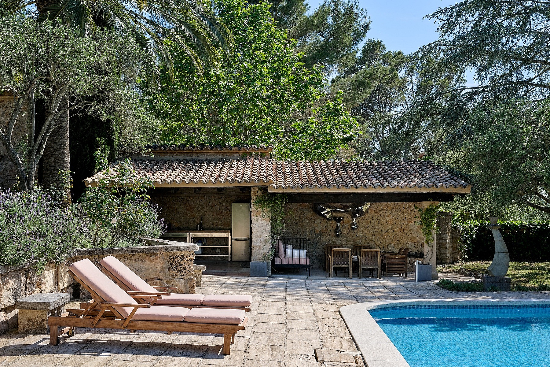 A stone-paved patio with two wooden sun loungers beside a swimming pool, featuring a covered outdoor dining area with a stone wall and rustic tiled roof.