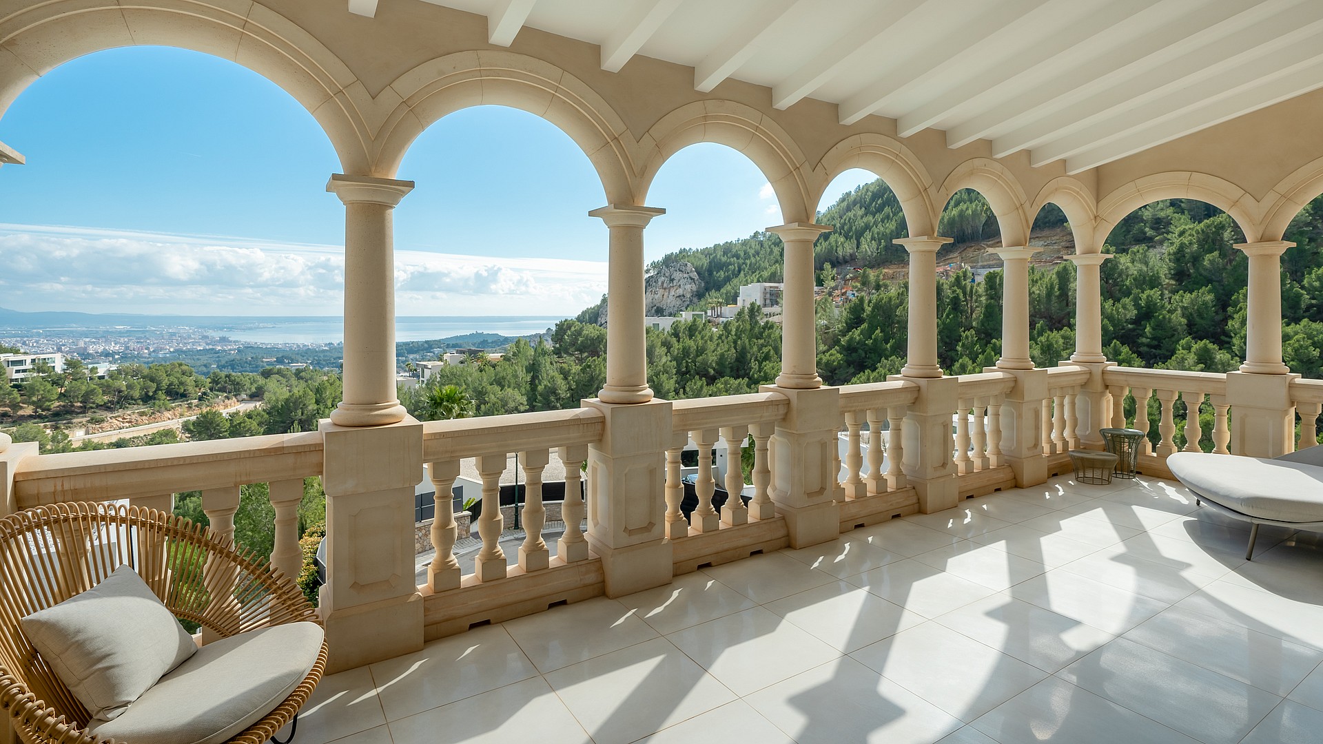 A covered stone loggia with arched columns and a balustrade overlooking a green valley and distant coastal city under clear skies.