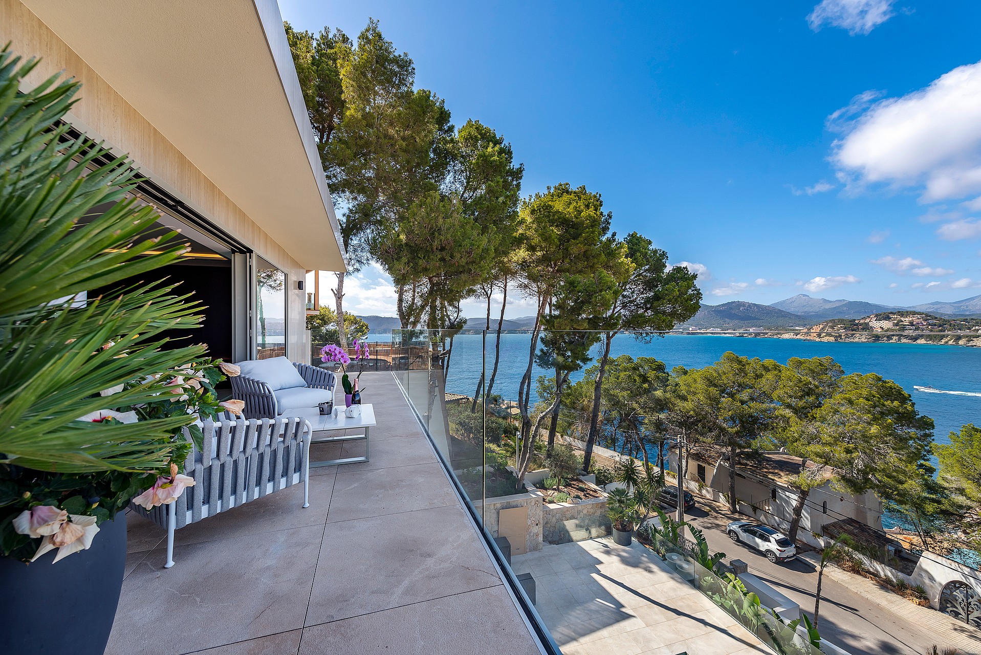 A wide terrace with grey outdoor furniture, glass railings, and a view overlooking the Mediterranean coastline and pines.