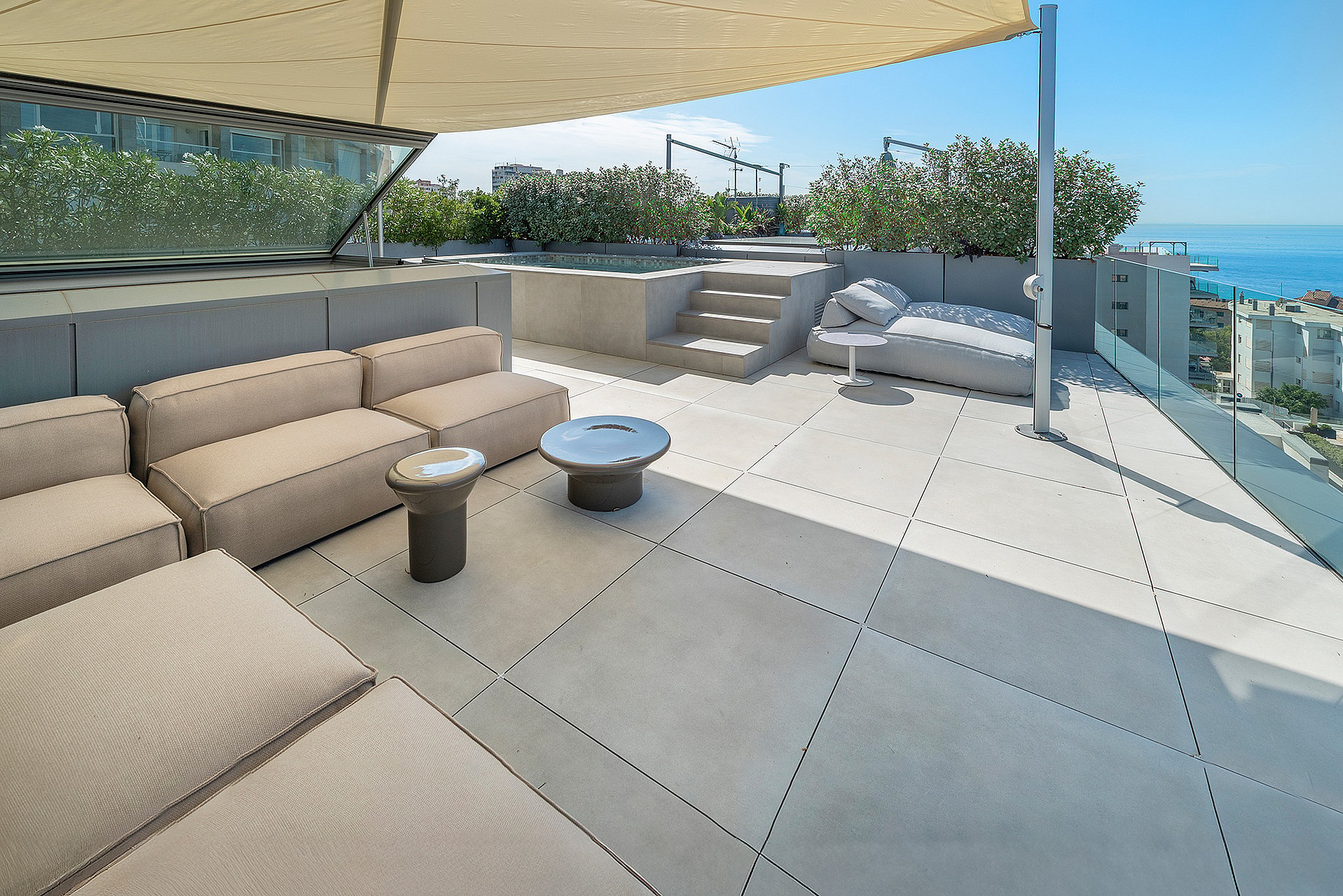 An expansive rooftop terrace with modular cream-colored seating, a glass balustrade, and a view overlooking the Mediterranean.