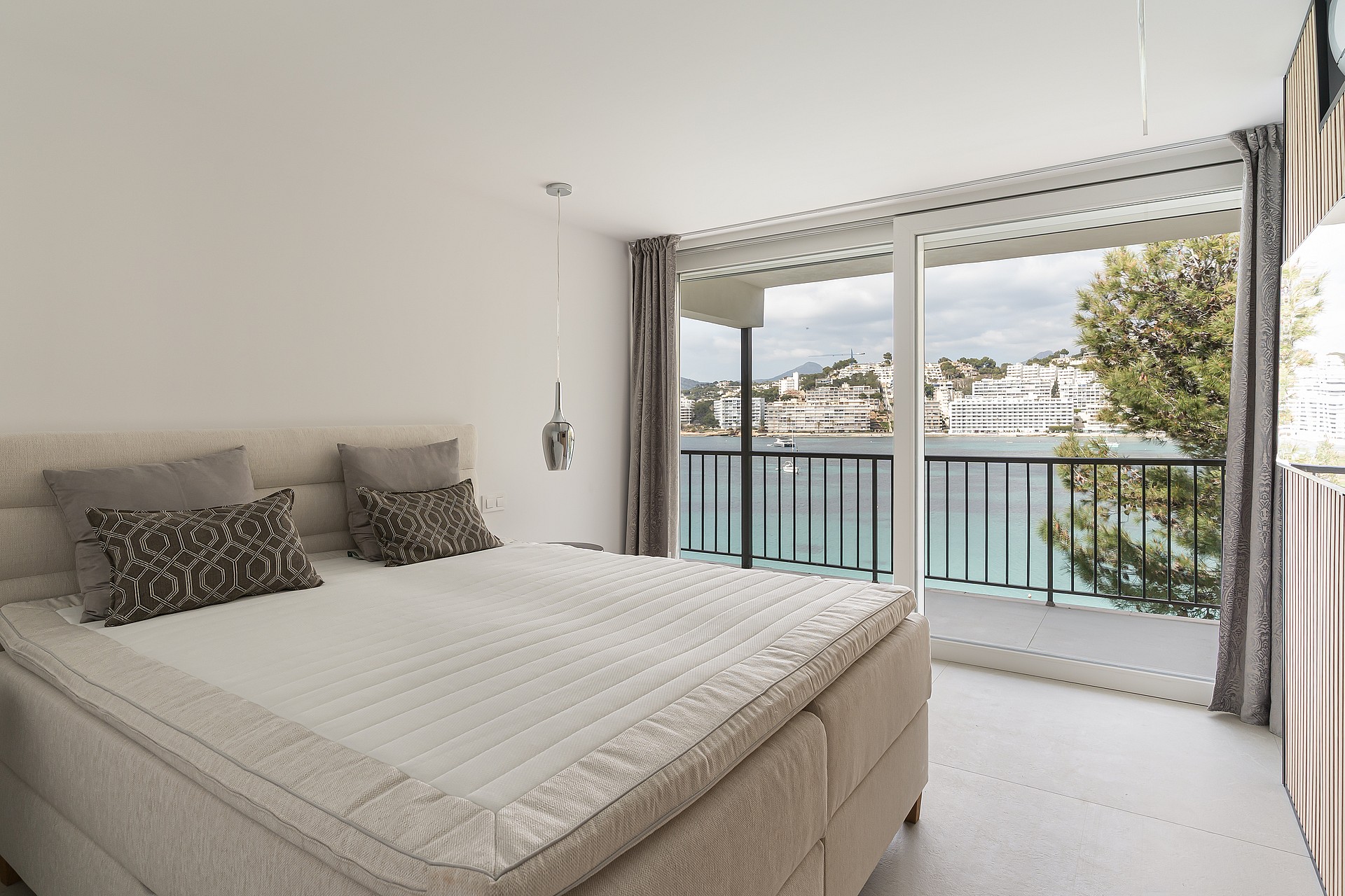 Modern master bedroom featuring a large sliding glass door opening to a private balcony with sea and town views.
