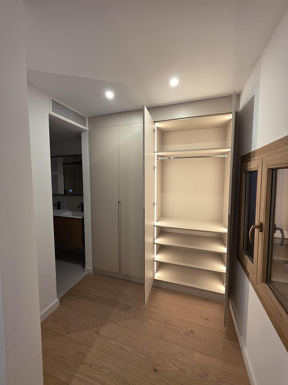A modern dressing area featuring floor-to-ceiling built-in cabinetry with integrated LED lighting and light oak hardwood floors.