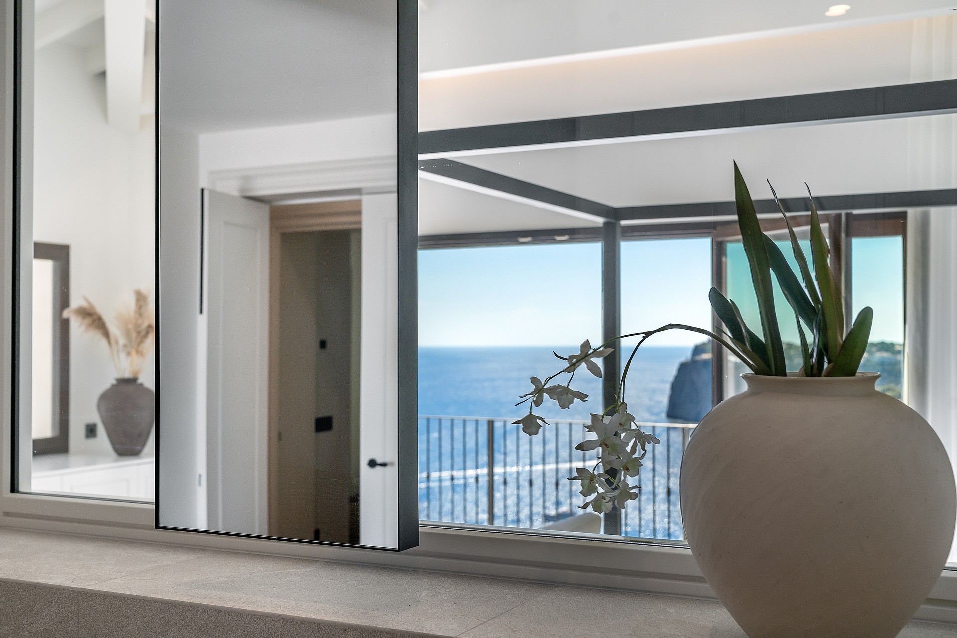 A clay vase with orchids reflects a metal bed frame and the blue sea.