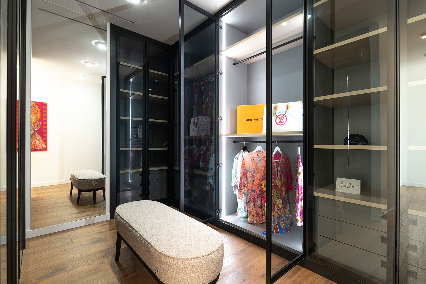 Walk-in closet with dark metal and glass cabinetry, integrated LED lighting, and a textured ottoman on hardwood flooring.