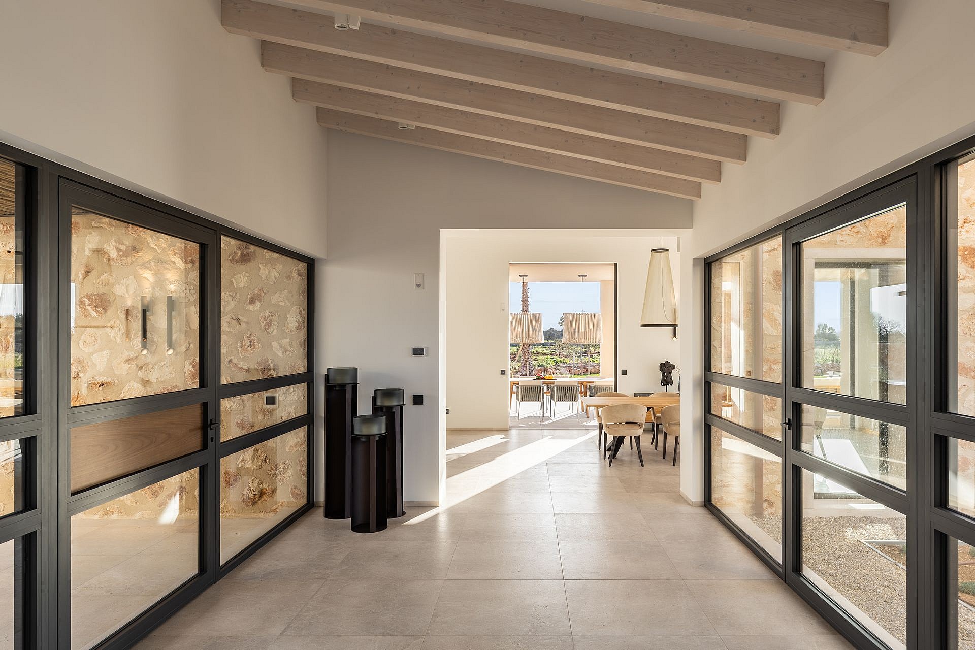 Interior hallway of a stone finca with large black-framed glass doors, light stone floors, and high wood-beamed ceilings.