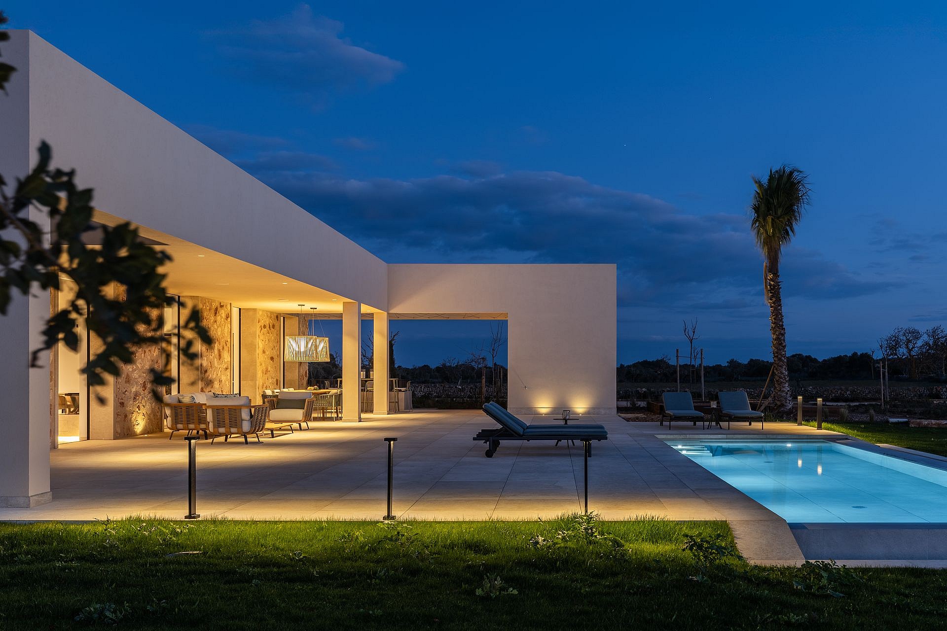 Exterior view of a modern finca at twilight, featuring a lit swimming pool, stone terrace, and warm architectural lighting.