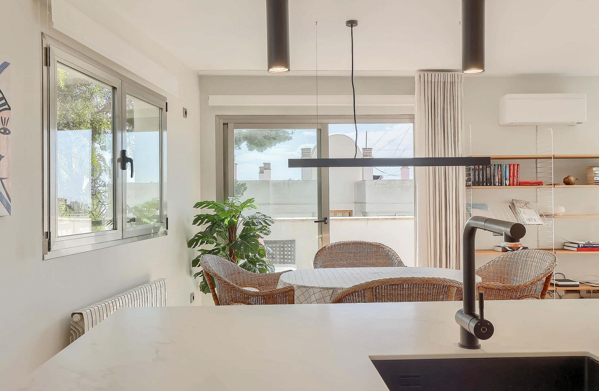 A modern kitchen foreground with a white countertop and dark faucet, opening into a dining area with wicker chairs and terrace access.