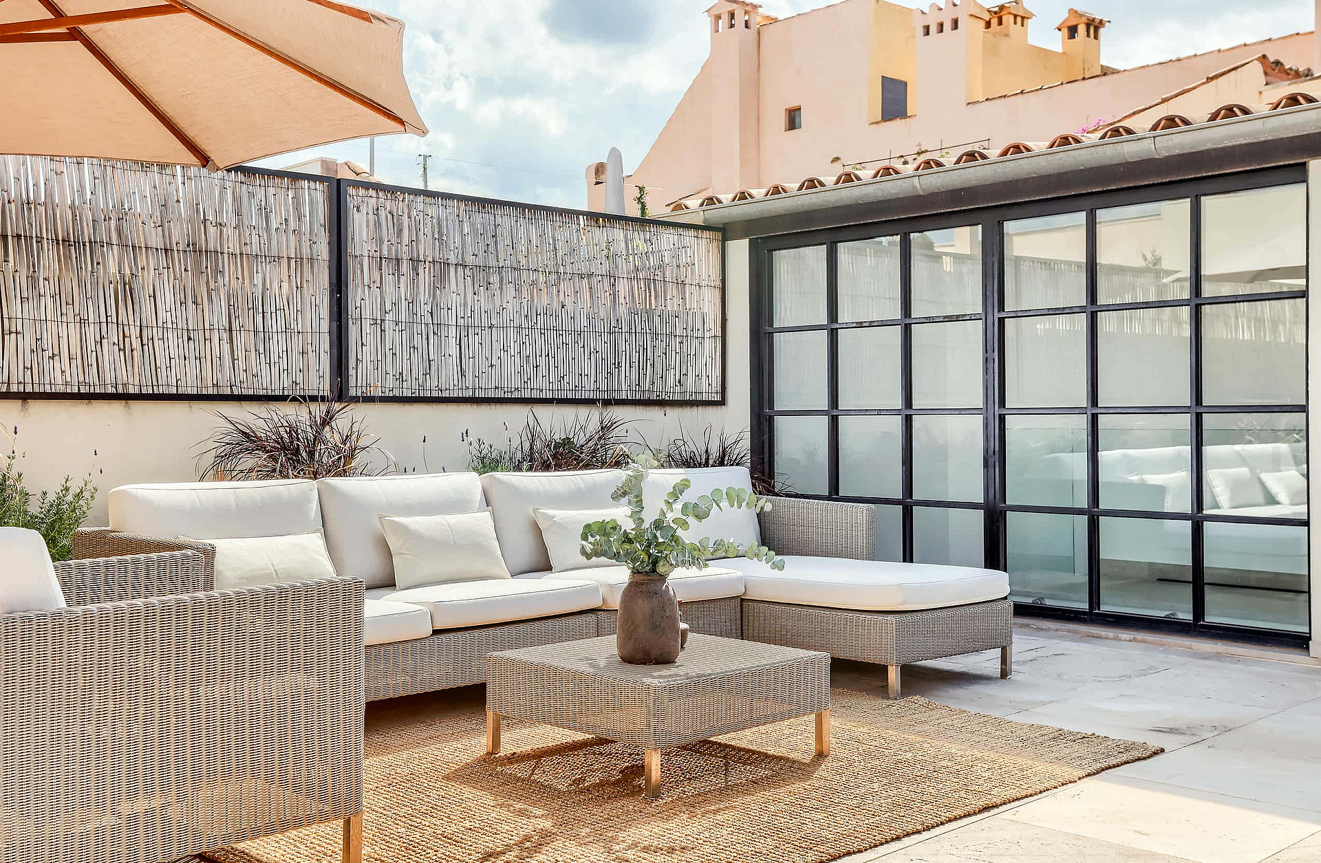 A private outdoor terrace with woven beige sofa sets, a coffee table on a natural fiber rug, and black steel-framed glass doors.