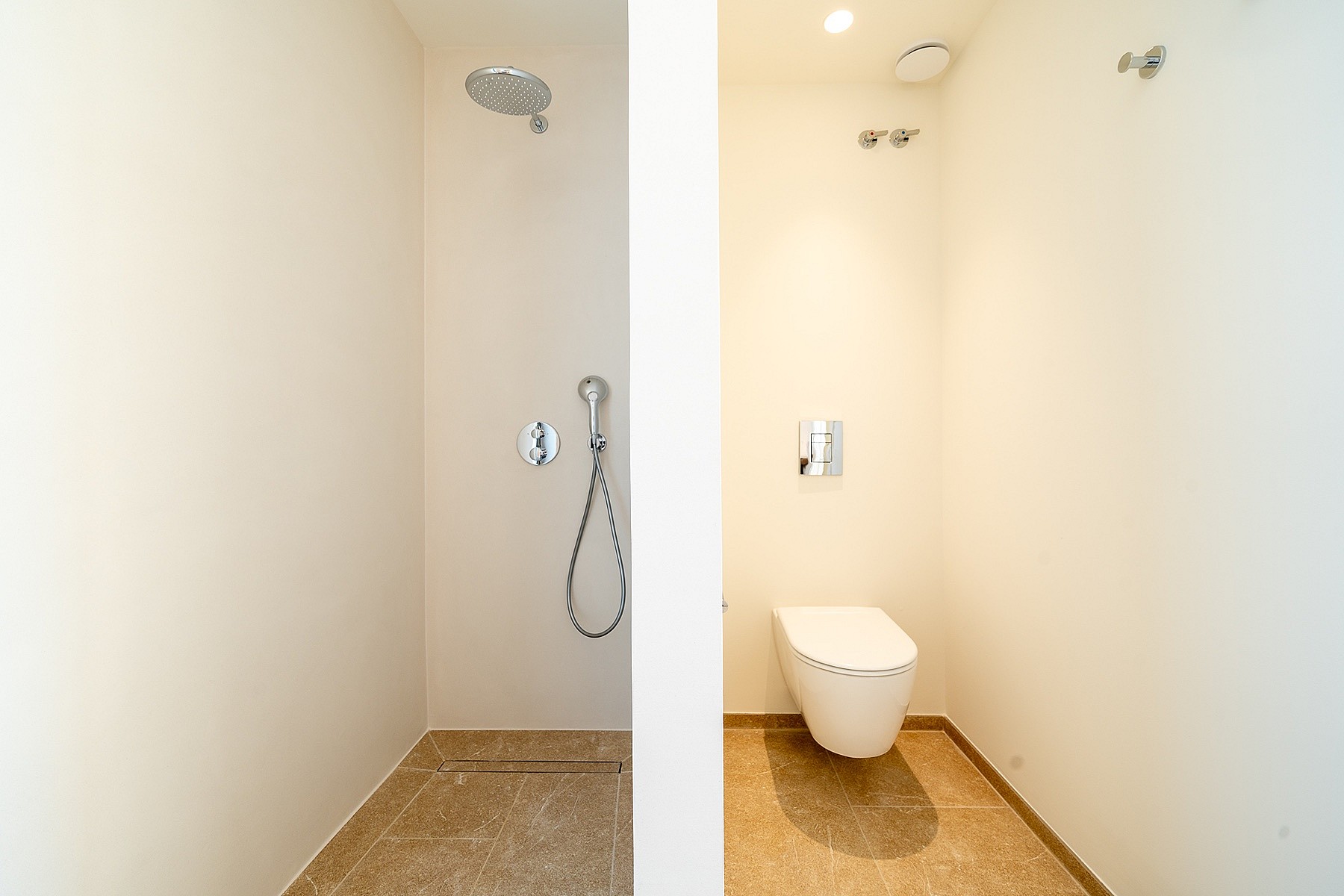 A minimalist bathroom section featuring a wall-mounted toilet and a walk-in shower with chrome fixtures and tiled floors.