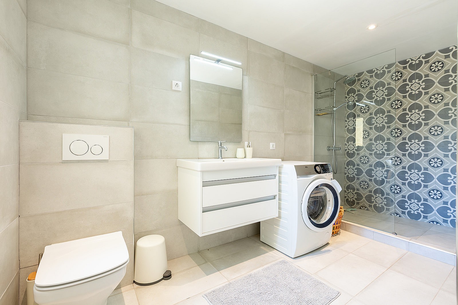 A modern bathroom featuring large light-grey wall tiles, a white vanity, a wall-mounted toilet, and a glass-enclosed shower with patterned floral tiles.