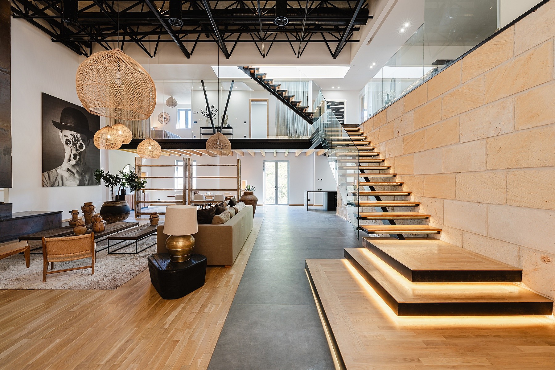 Double-height living room featuring a floating wooden staircase, limestone feature wall, and industrial black steel ceiling trusses.