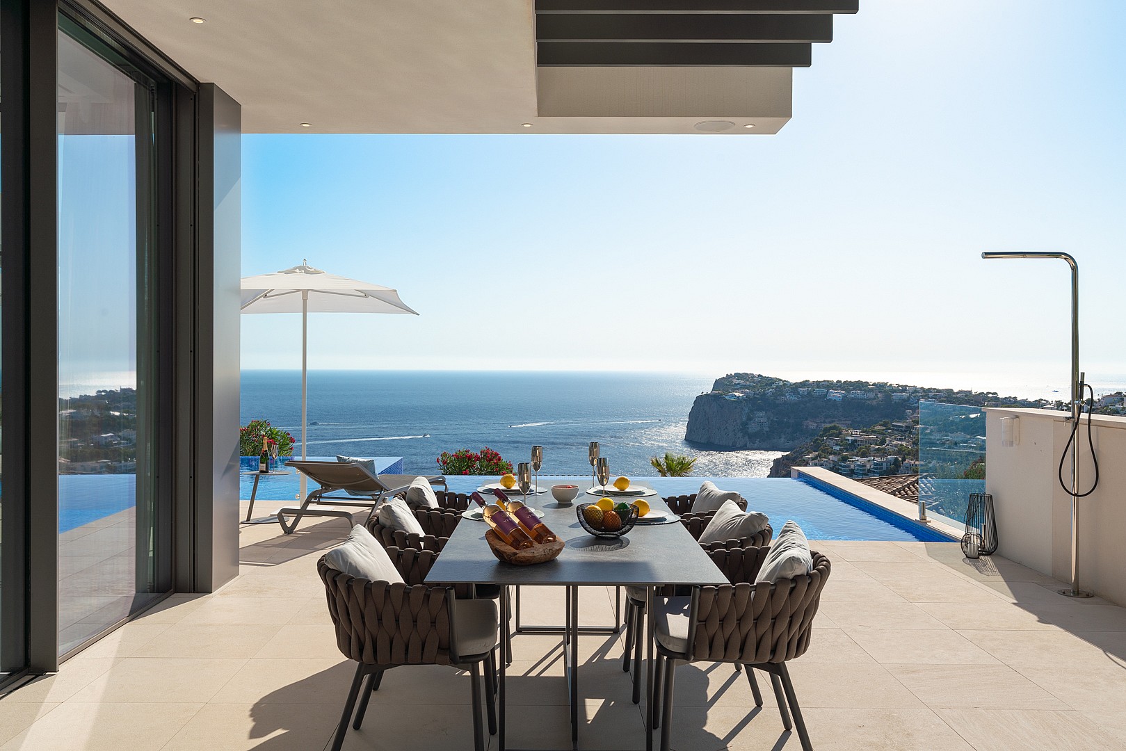 Covered outdoor dining terrace with a dark metal table, woven chairs, and an infinity pool edge against a Mediterranean sea view.
